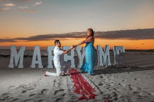 People pose with large white letters spelling 'MARRIAGE' on a sandy beach at sunset.