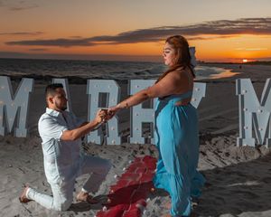 A romantic beach proposal at sunset with a blue dress and large decorative letters spelling MARRY ME in the background.