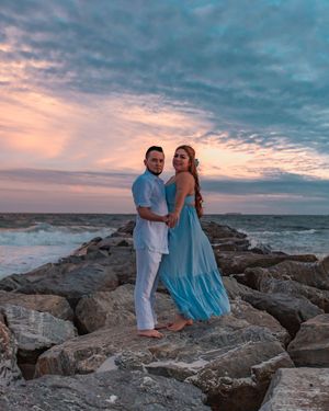 Couple embraces on rocky coastal shoreline at sunset with dramatic cloudy sky and ocean backdrop.