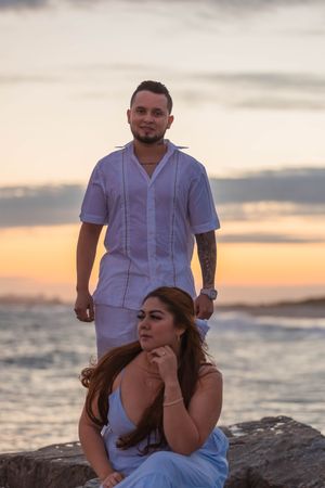 Couple in white outfits sitting on coastal rocks during sunset at the beach.