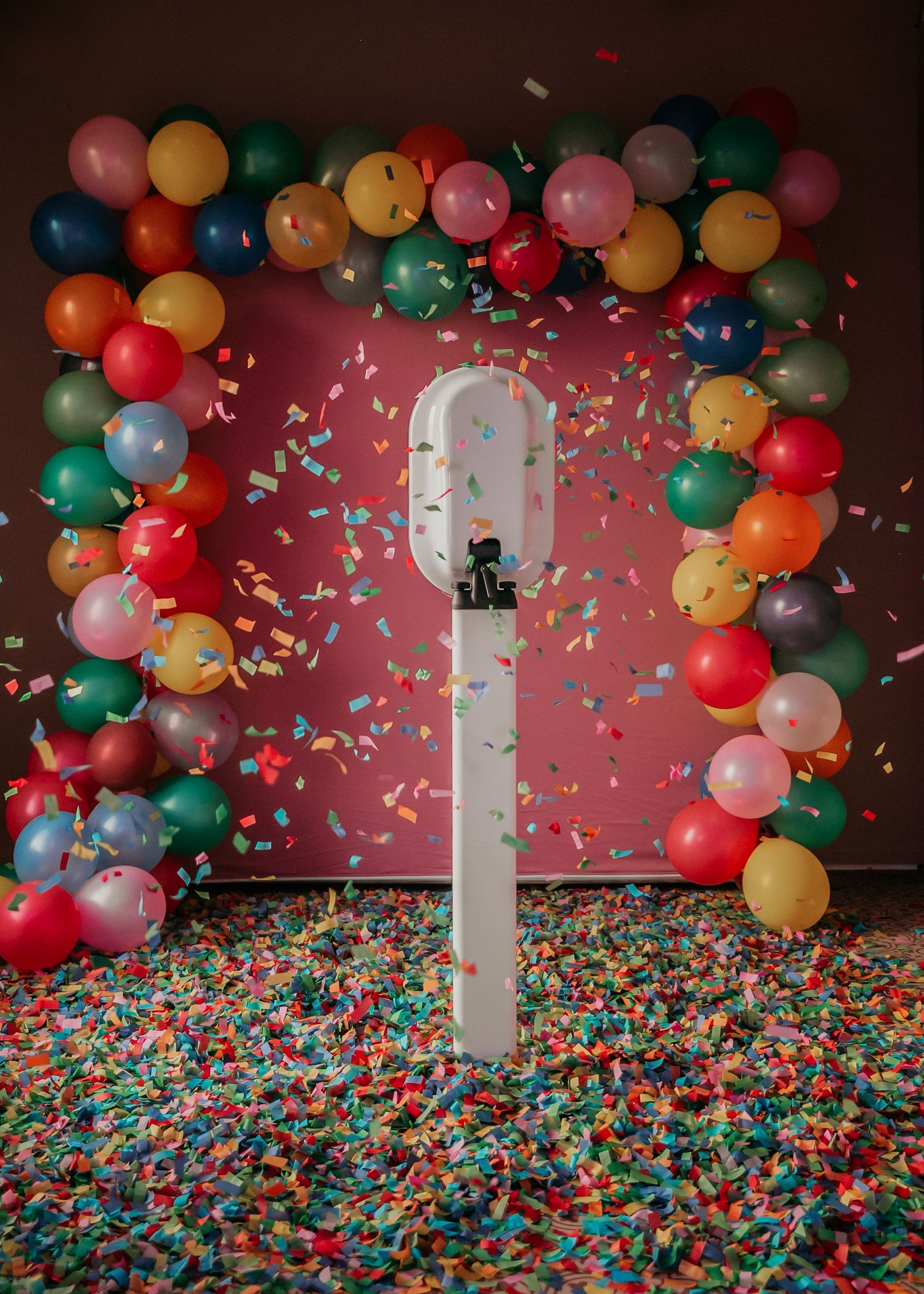 Colorful balloon arch and confetti create a festive party backdrop against a dark wall with a bright phone stand in center.