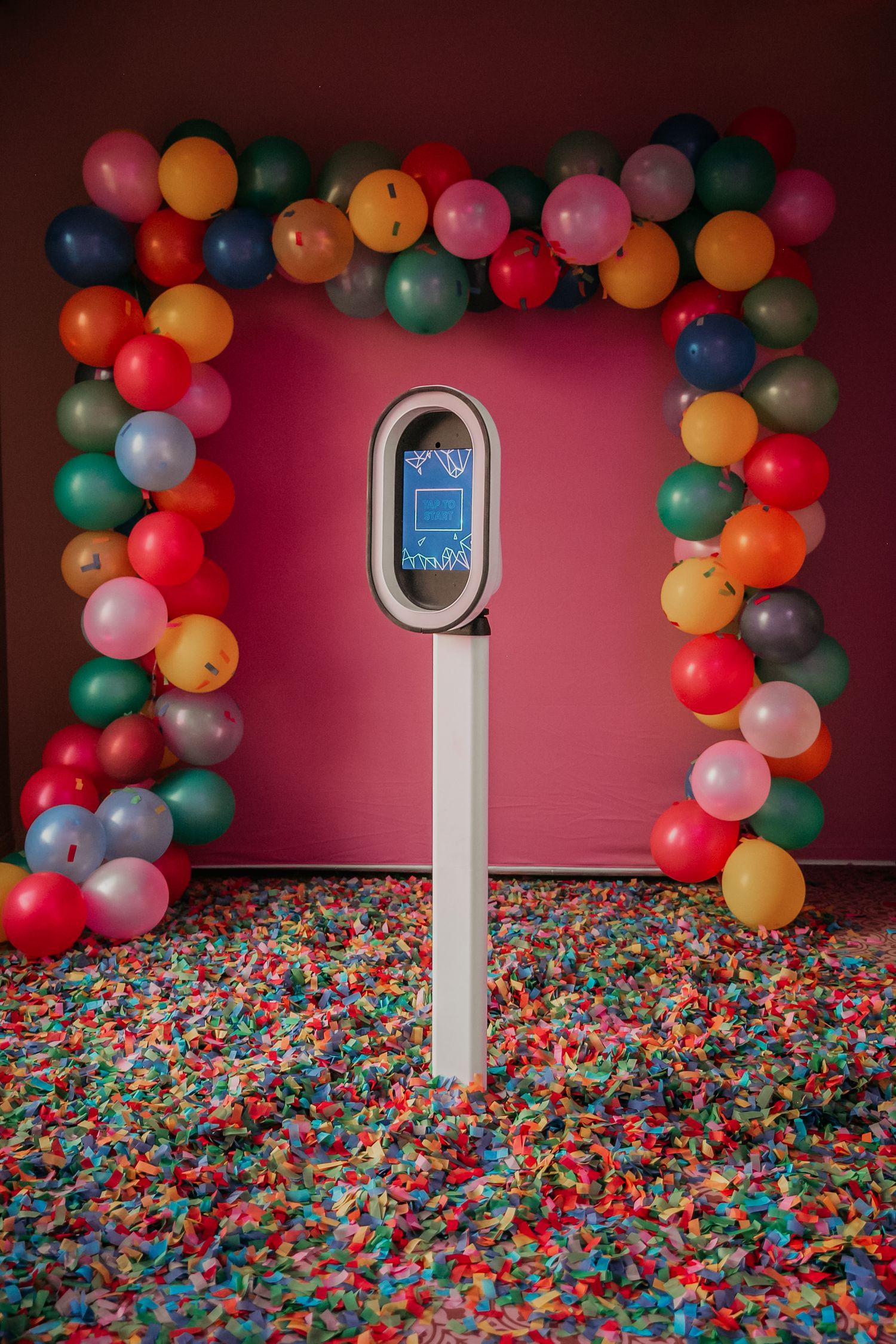 A white pedestal phone display stands amid a sea of colorful balloons and confetti against a red wall.