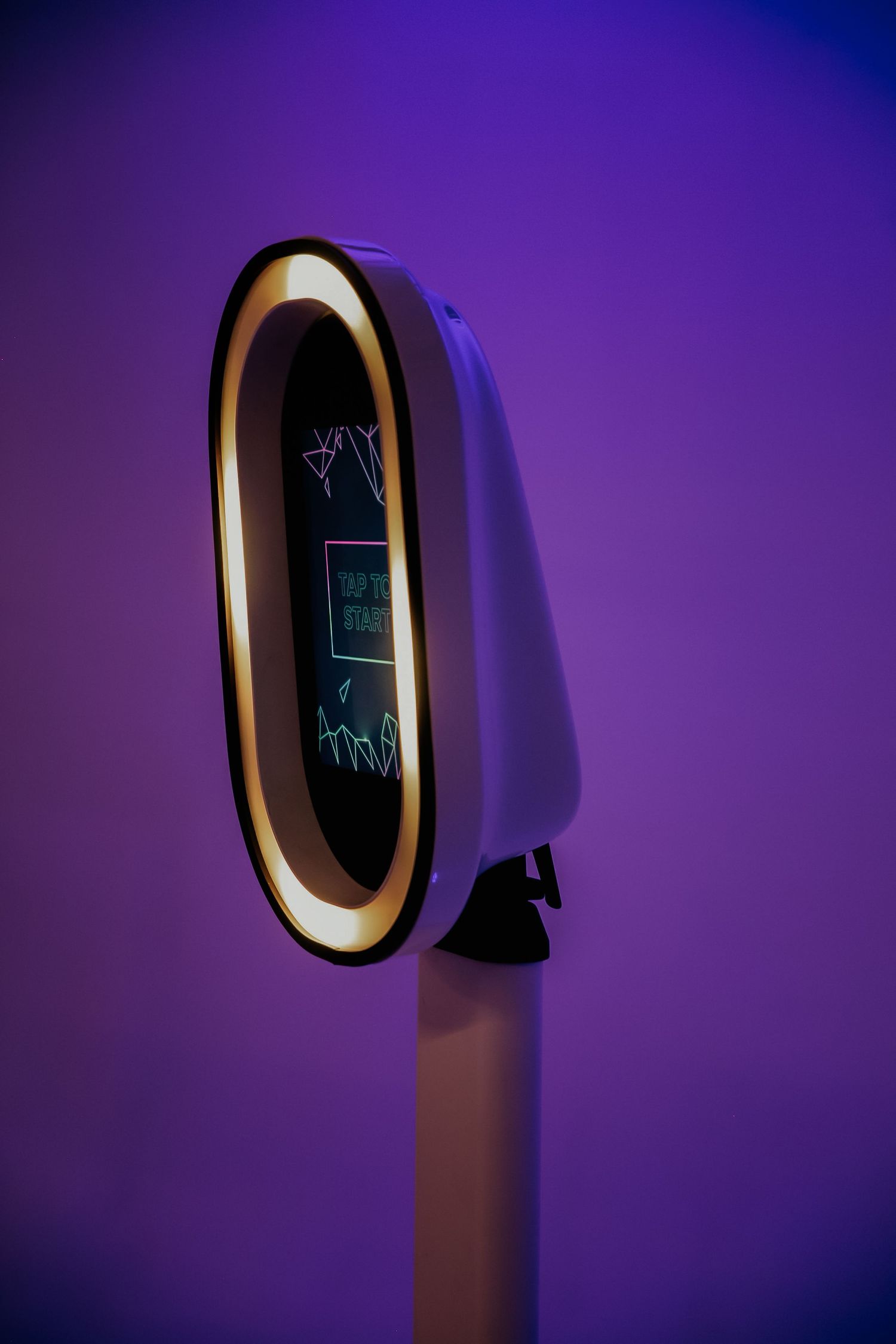 Modern photo booth with illuminated oval frame and digital screen display against purple gradient background.
