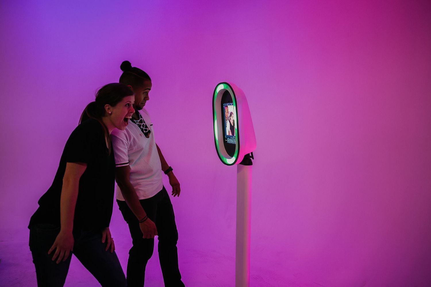 Two people walk past a photo booth kiosk lit by vibrant purple lighting against a pink background.