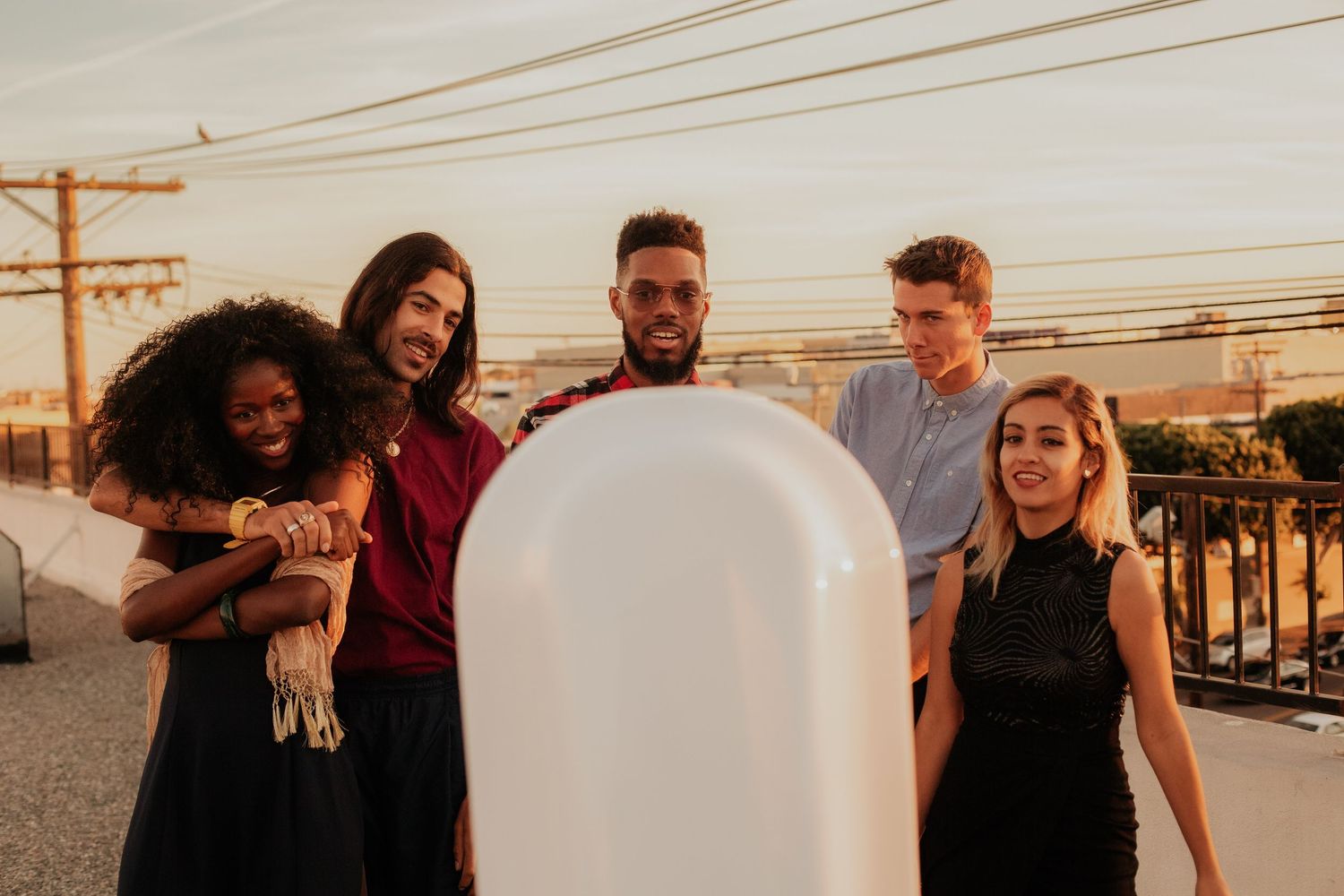 A diverse group of young people gathered outdoors at sunset around a white inflatable object in an urban setting.