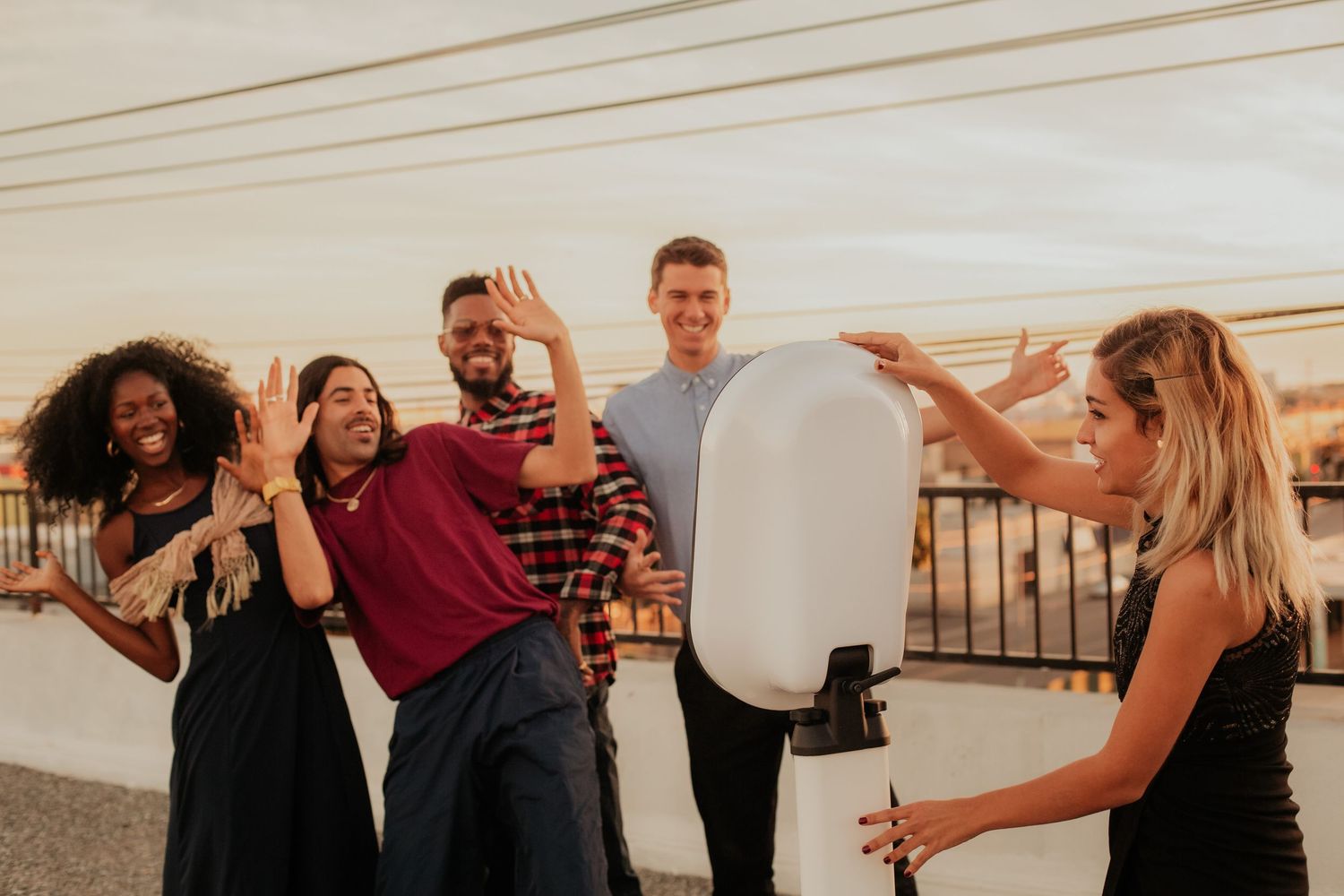 A group of friends laughing and posing together while someone adjusts a photography light on a balcony at sunset.