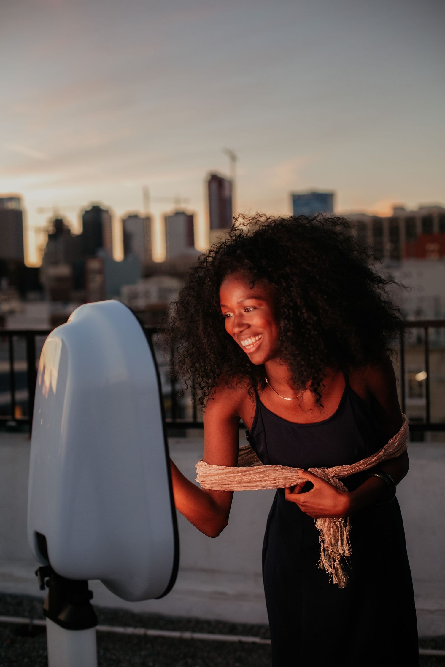 Person in a black dress reading a book next to a glowing white modern sculpture during sunset with city skyline in background.