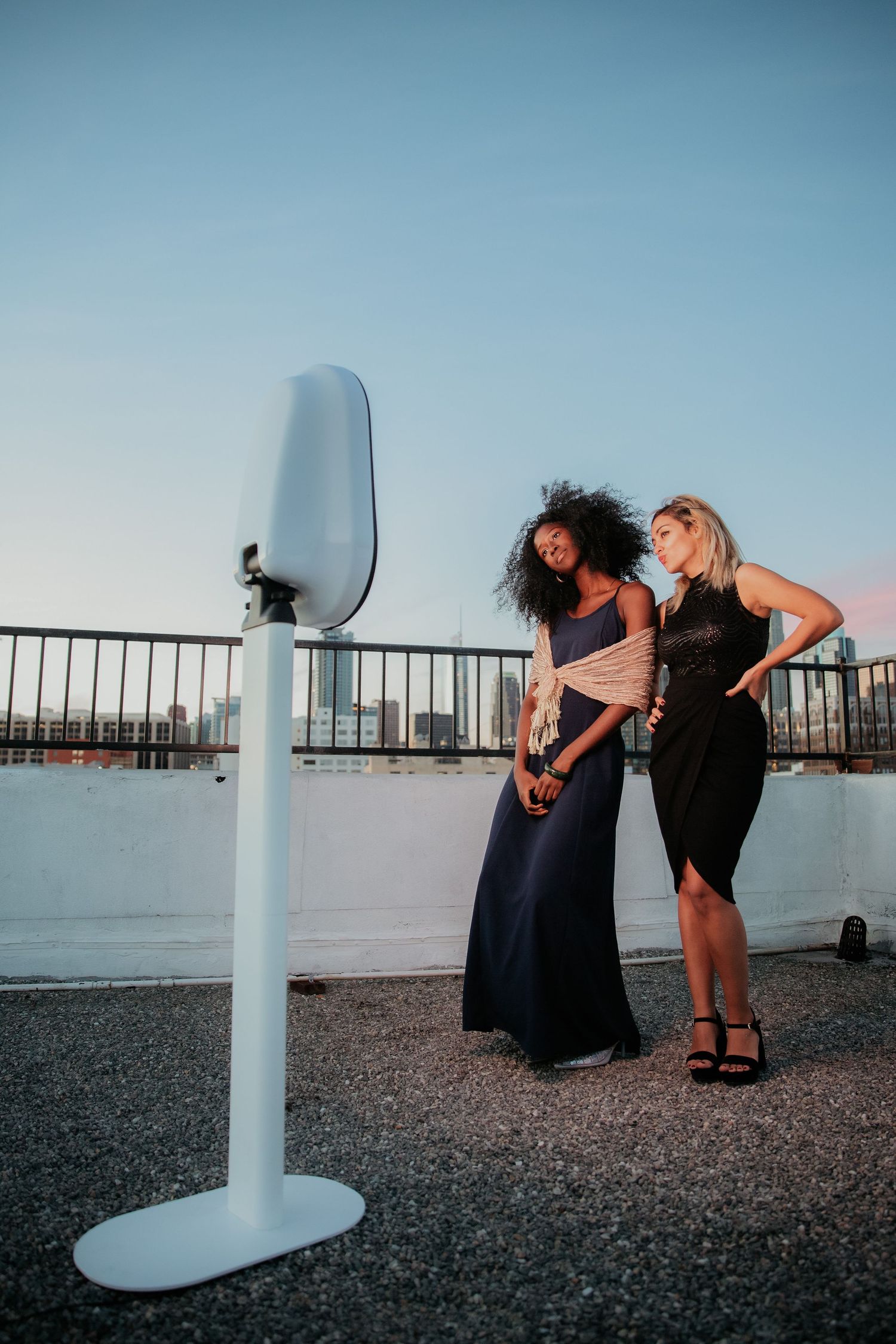 Two women in elegant dresses pose playfully next to a viewing binocular stand on a rooftop observation deck at sunset.