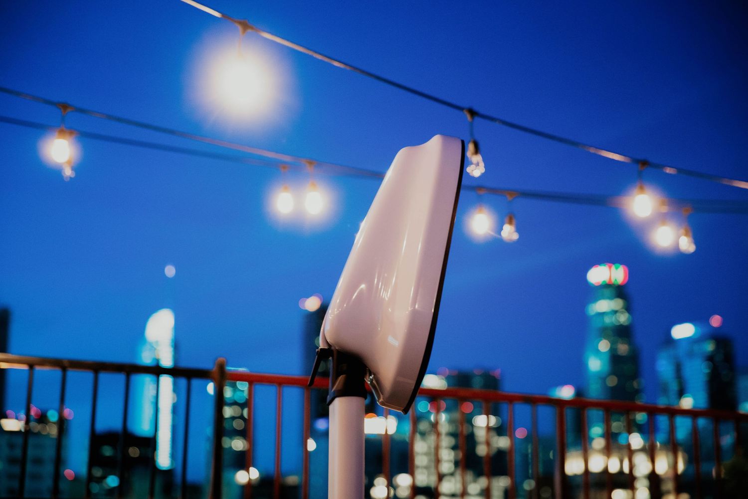 String lights illuminate a modern rooftop terrace with city lights and an outdoor television display in the evening.