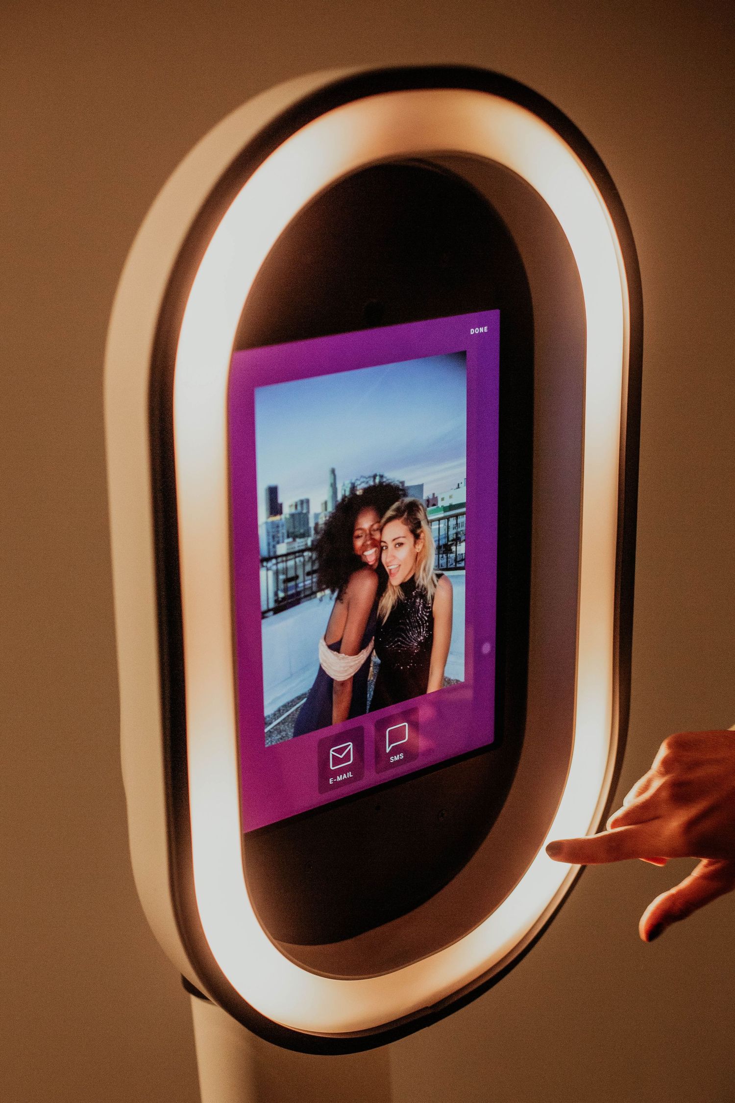 A photo booth with illuminated oval ring light displays a digital image with pink frame against a brown background.