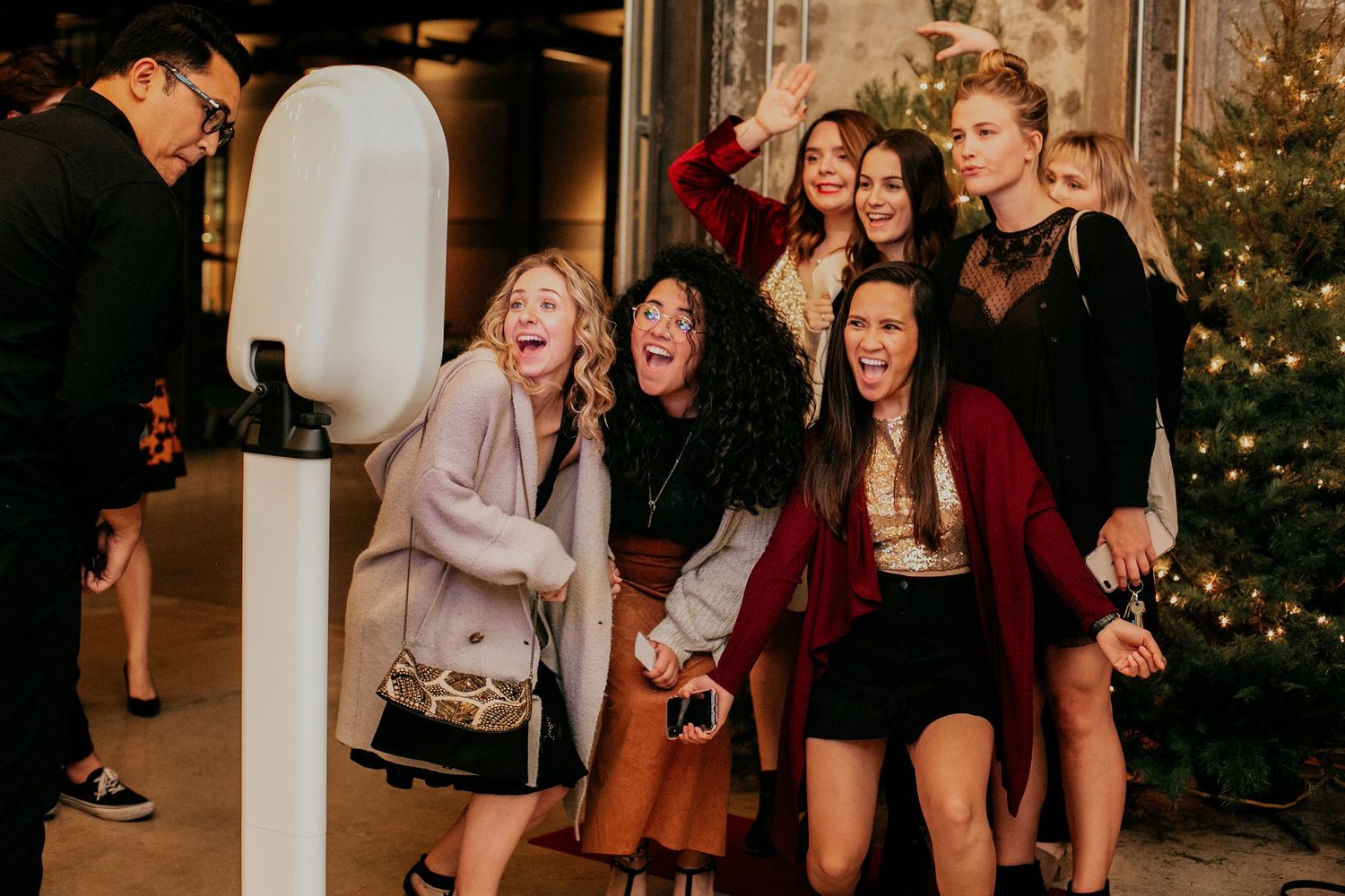 A group of casually dressed friends sharing laughter together during a nighttime holiday gathering near a Christmas tree.