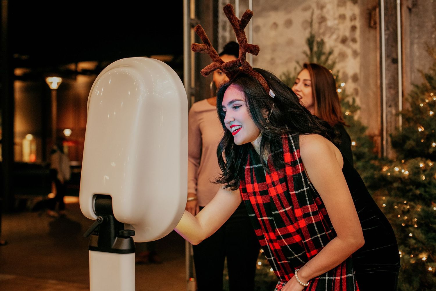 Person wearing plaid shirt interacting with white charging station or kiosk outdoors at night with holiday lights.