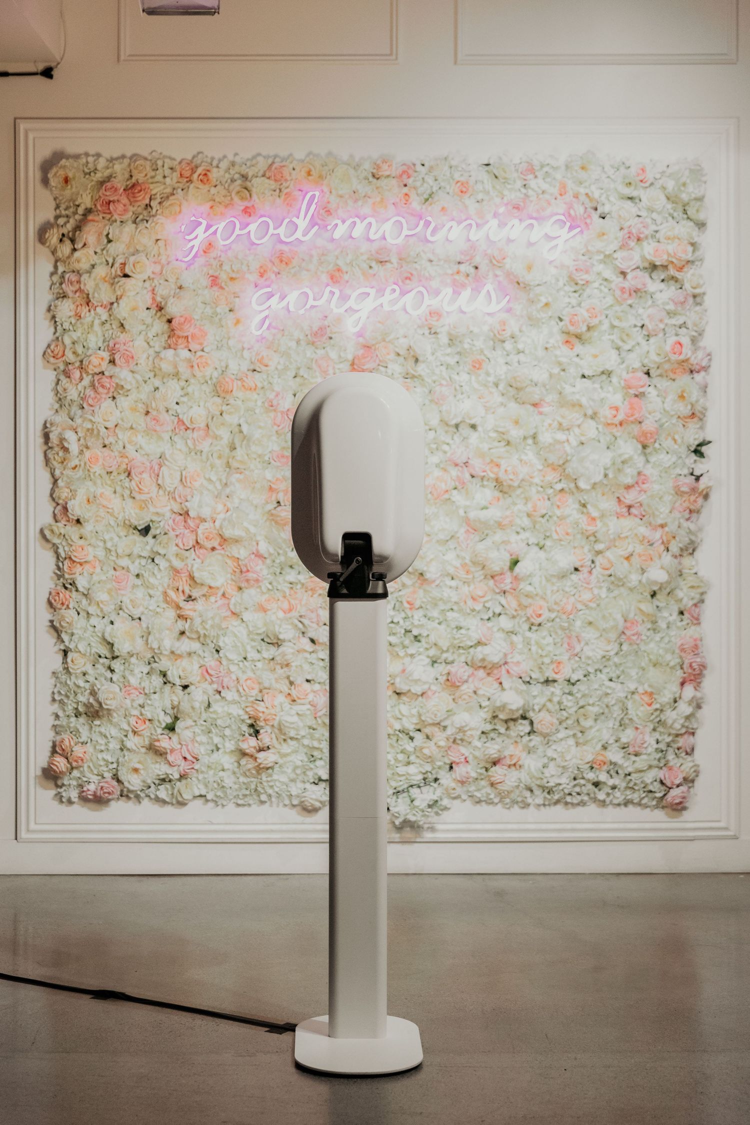 A white selfie station stand against a floral wall backdrop with pink neon sign reading 'good morning gorgeous'.