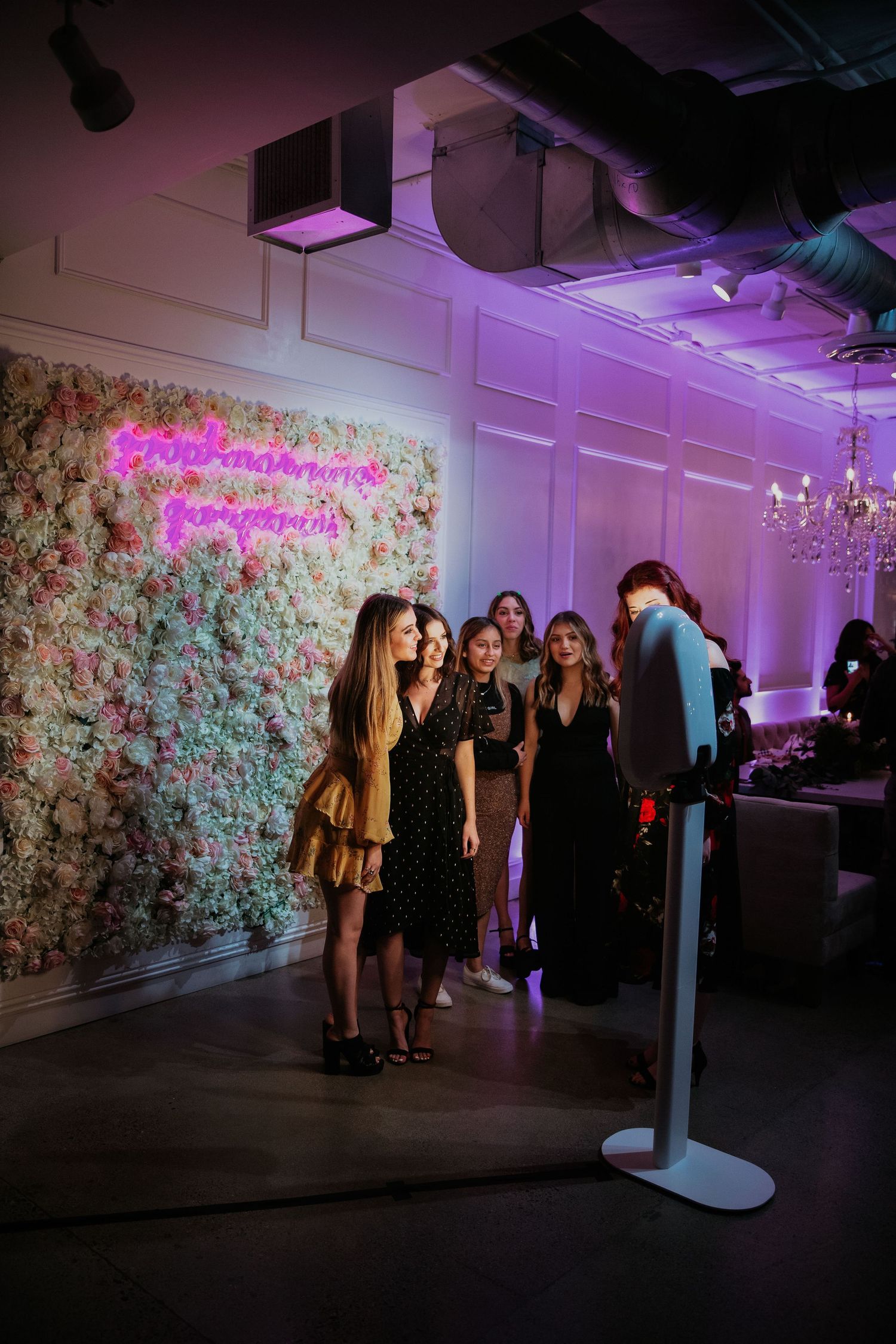 A group in formal attire poses in front of a floral wall backdrop with purple mood lighting at an elegant event venue.