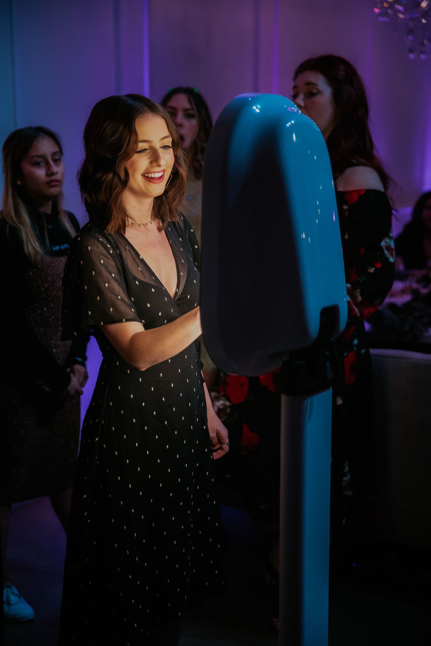 Woman in black sparkly evening dress smiles at blue neon-lit display at nighttime event.