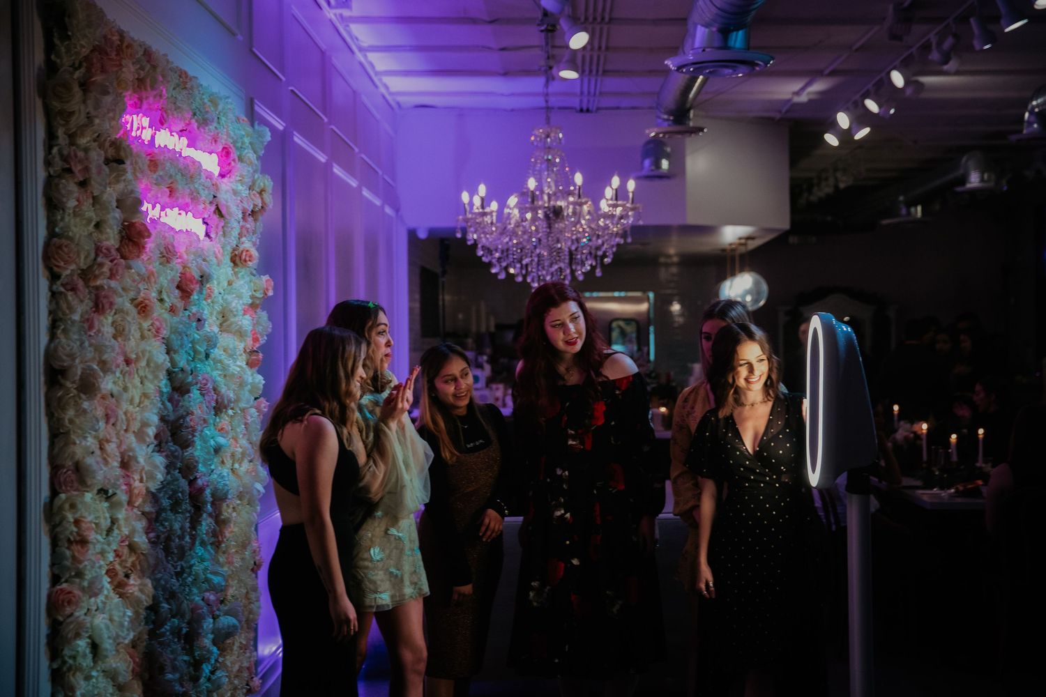 A dimly lit party venue with purple lighting, neon sign, and crystal chandelier creating an elegant nightclub atmosphere.