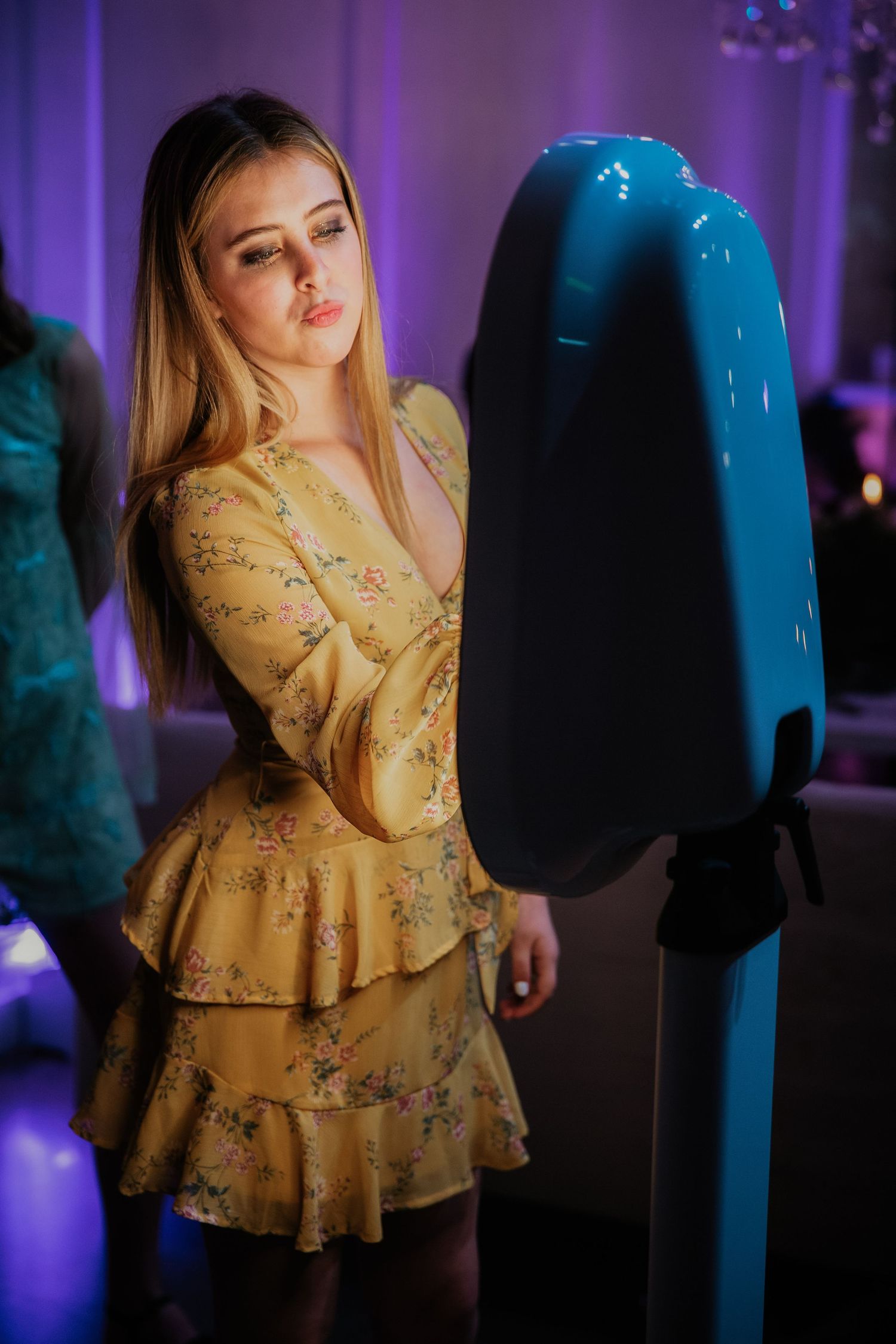 Person in yellow floral tiered dress posing next to a dark electronic device in purple-lit environment.