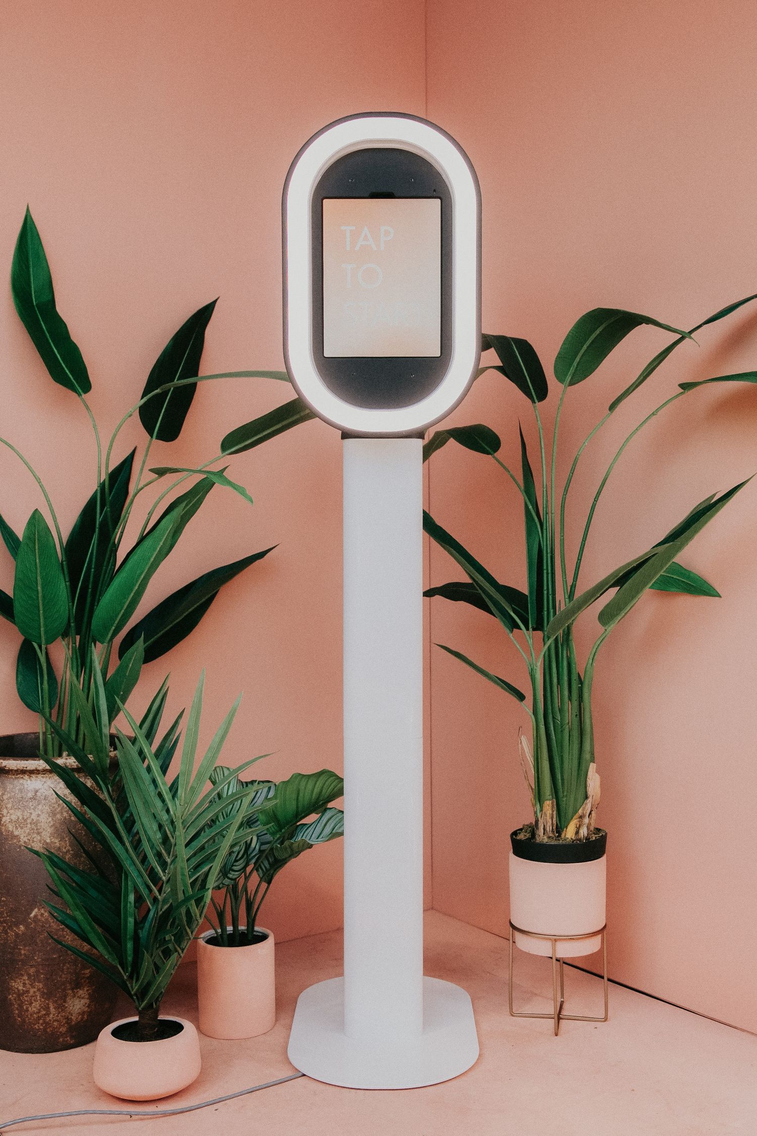 Modern white stand-alone selfie booth with digital screen display surrounded by potted plants against pink wall.