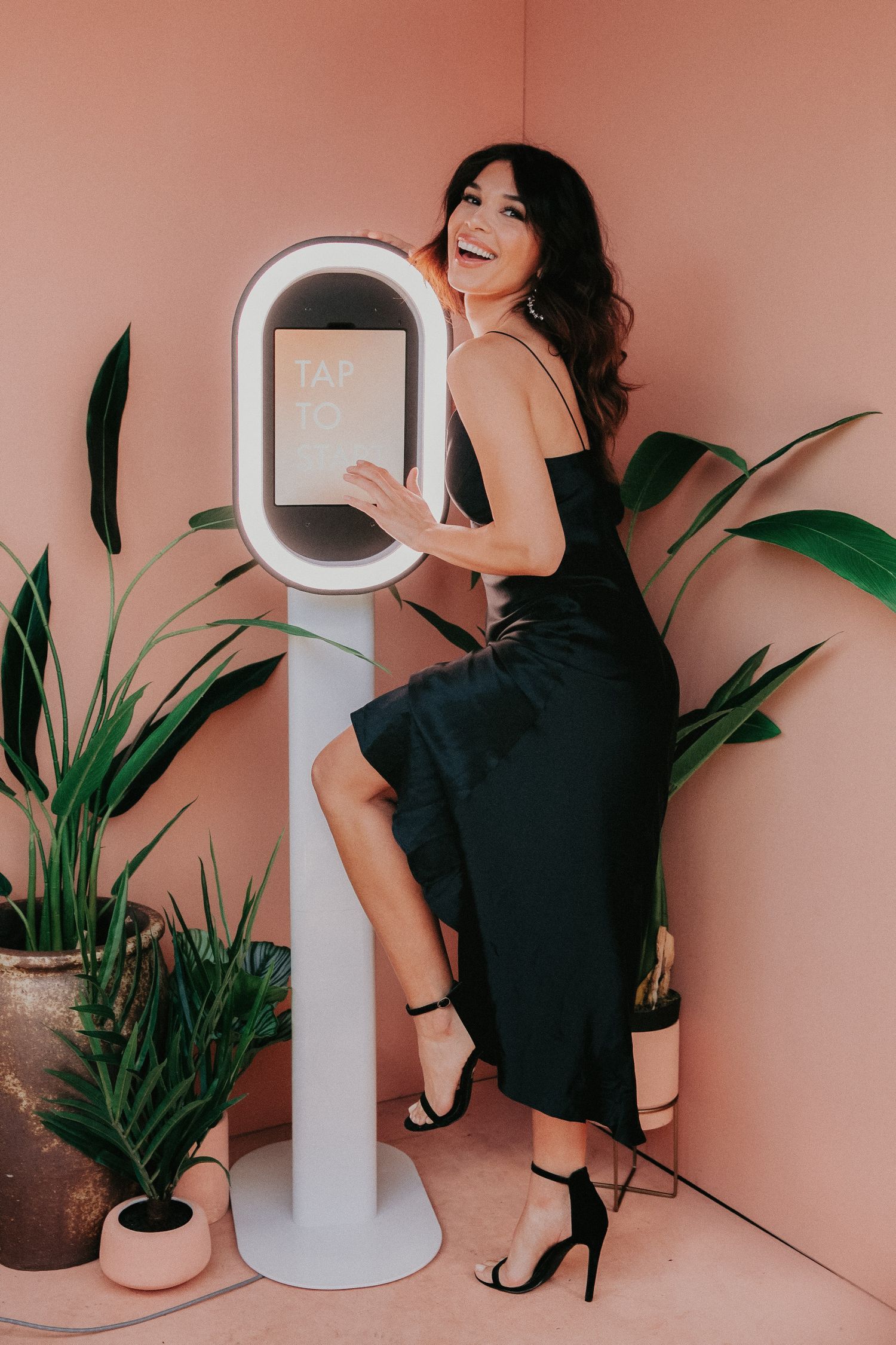A woman in a black dress poses in a series of four artistic photos with tropical plants against a pink backdrop.