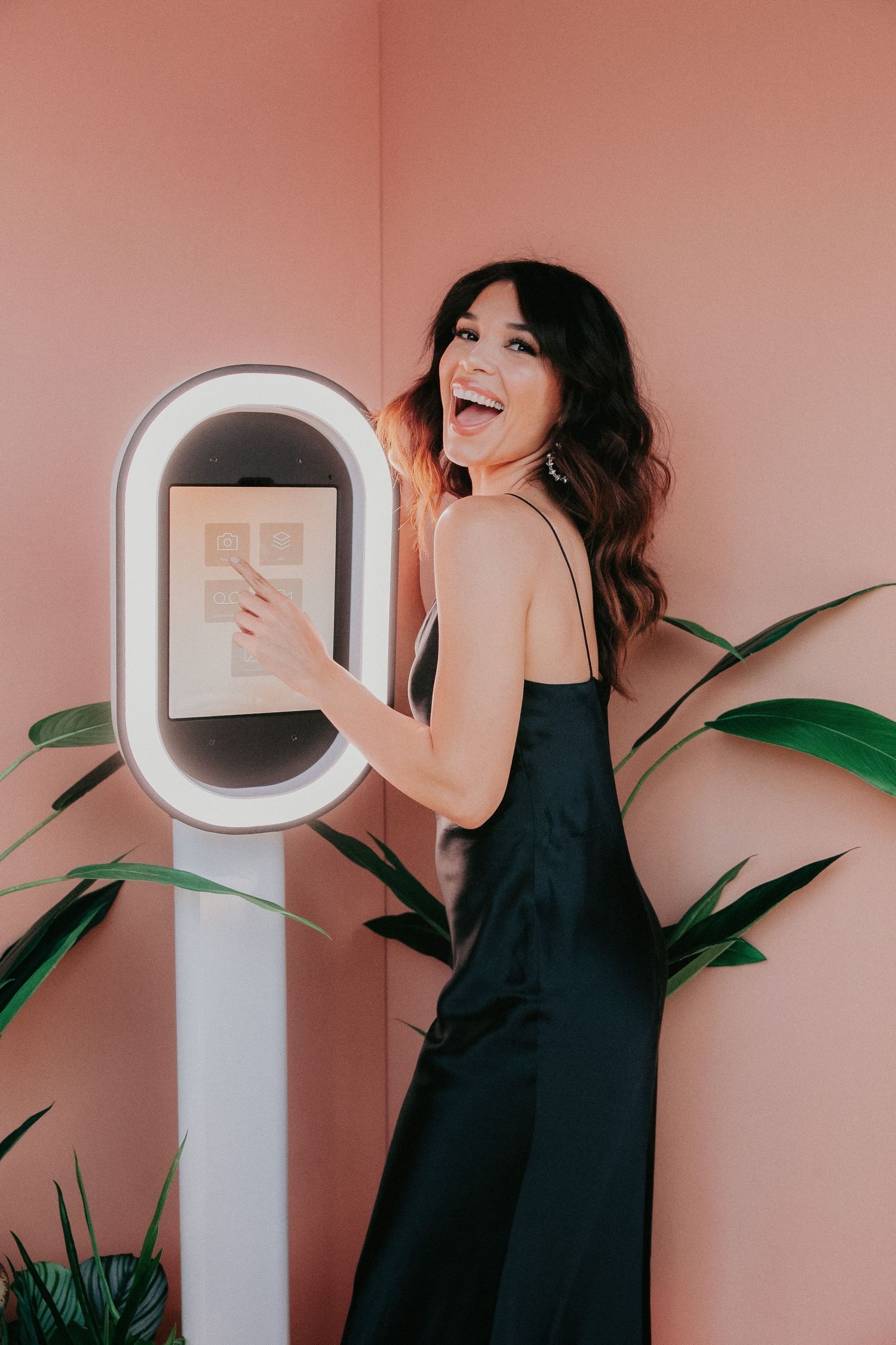 Person in black jumpsuit posing with interactive touch screen display next to tropical plants against pink wall.