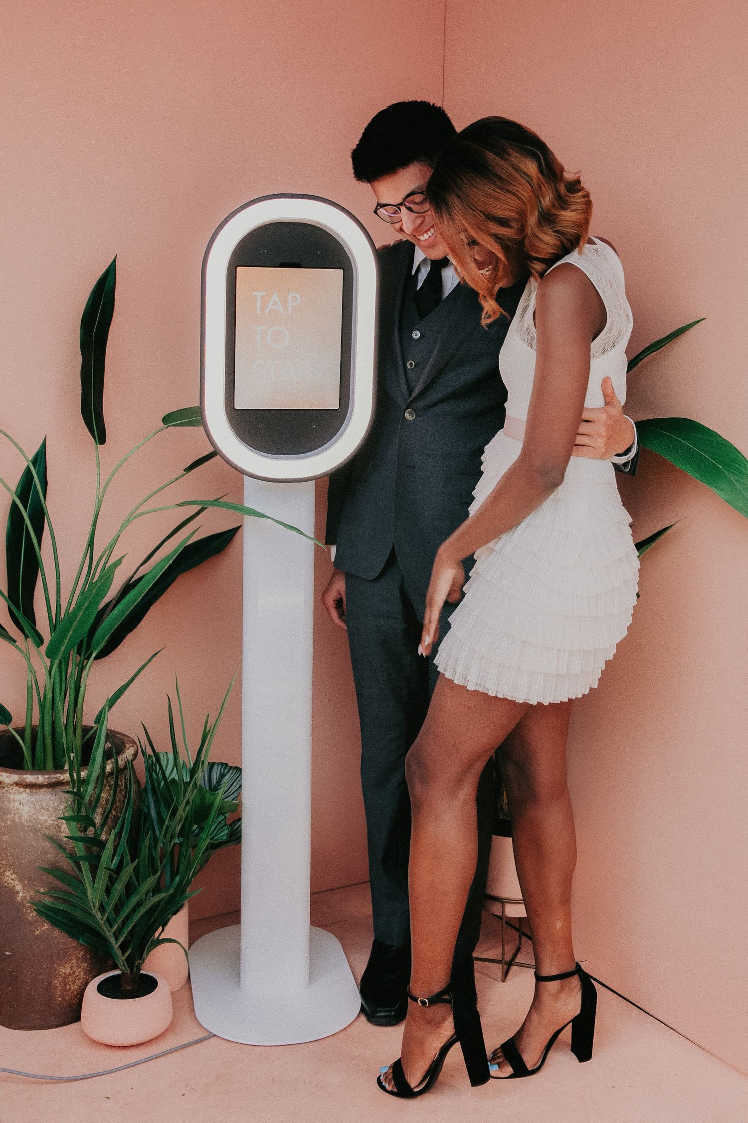 Couple in black and white outfits poses intimately against pink wall with decorative plants and mirror.