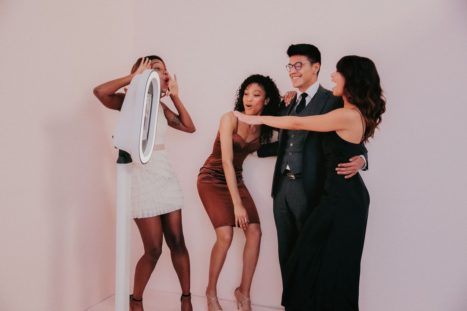Group of friends laughing and dancing together at a party in brown and black outfits against pink backdrop.