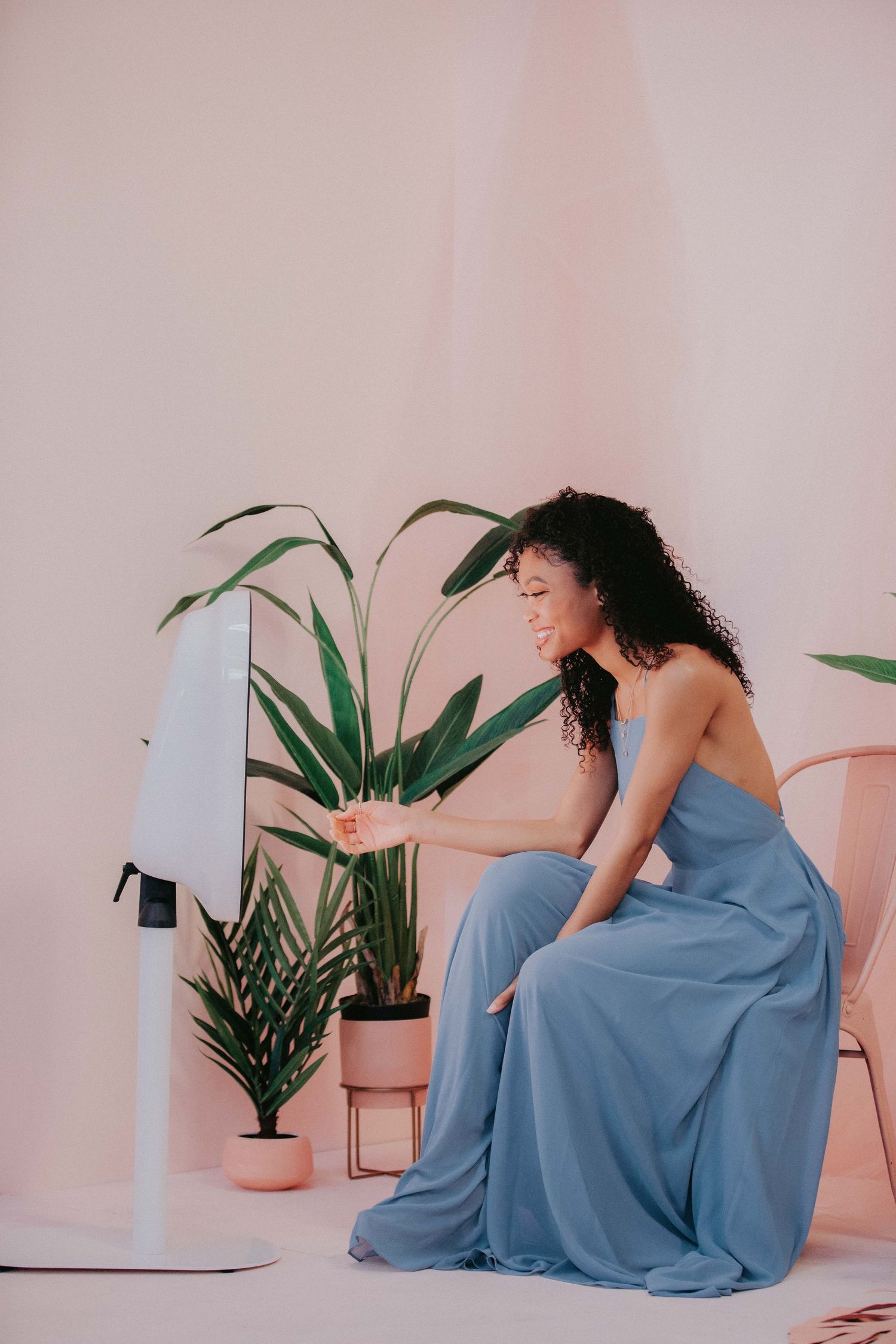 Woman in flowing blue dress sits elegantly beside potted plants against pink wall while interacting with device.