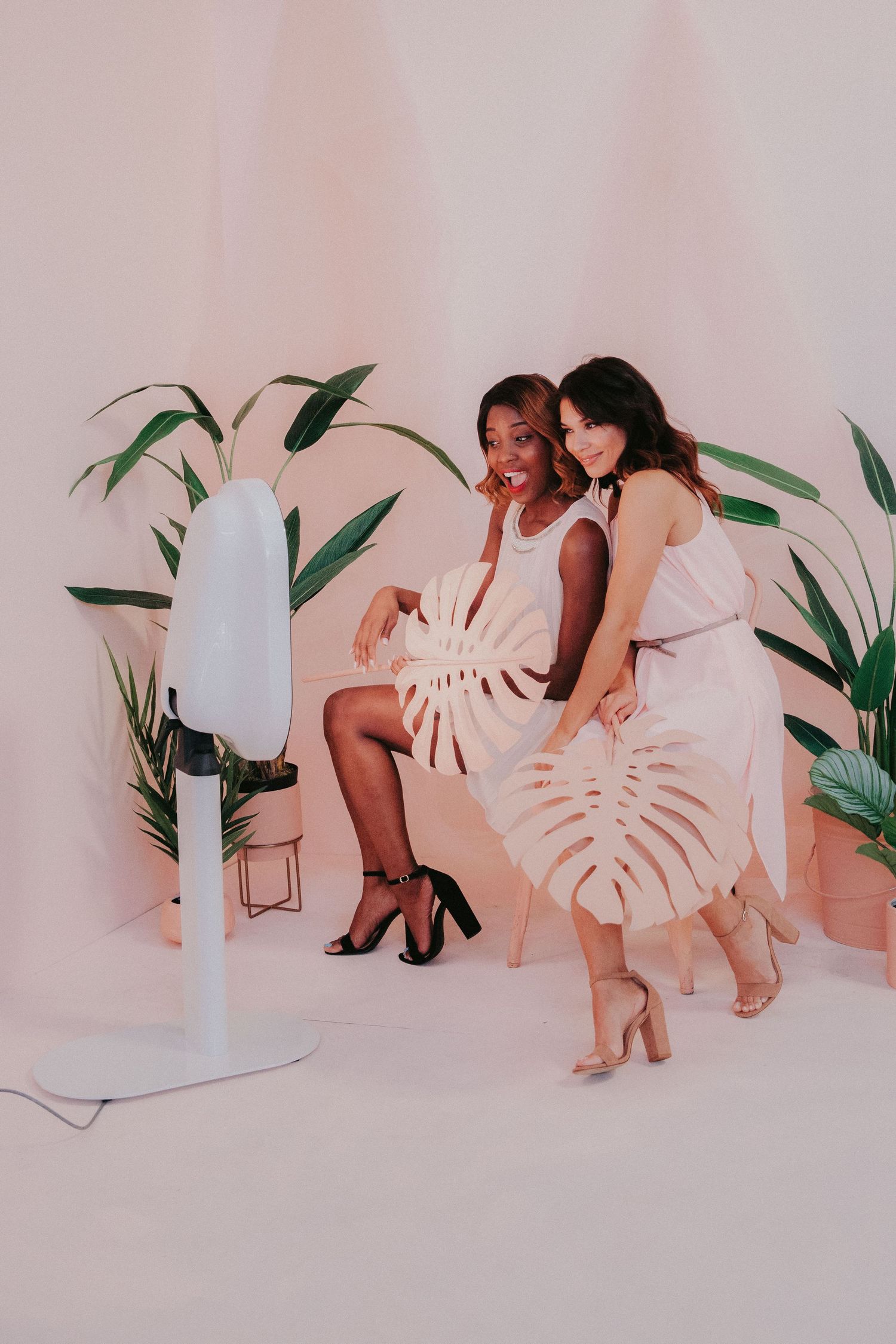Two friends in white dresses pose together in a studio with tropical plants and a floor lamp against a pink backdrop.