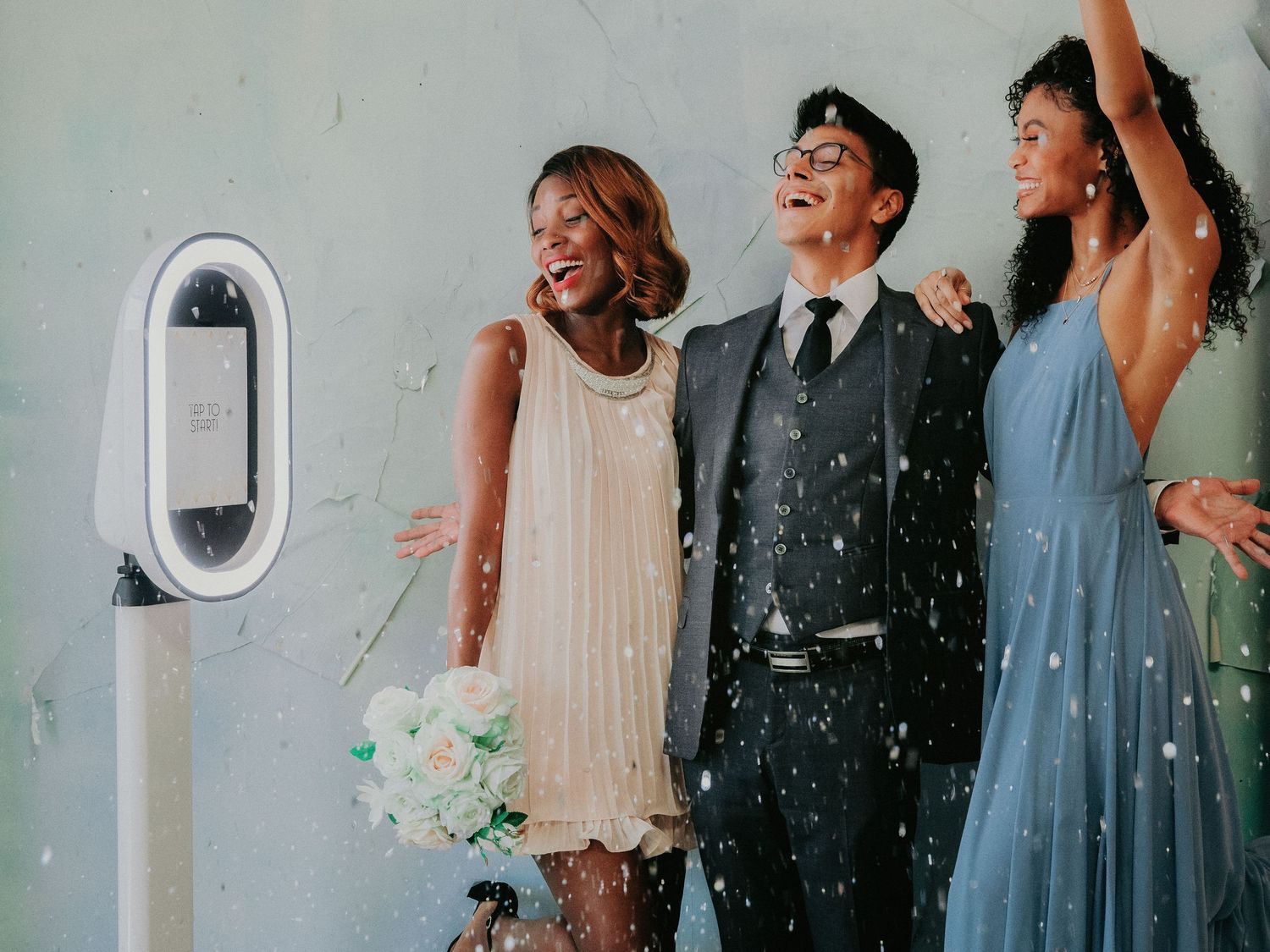 Three people in formal attire pose and laugh together next to a white wall-mounted control panel while confetti falls around them.