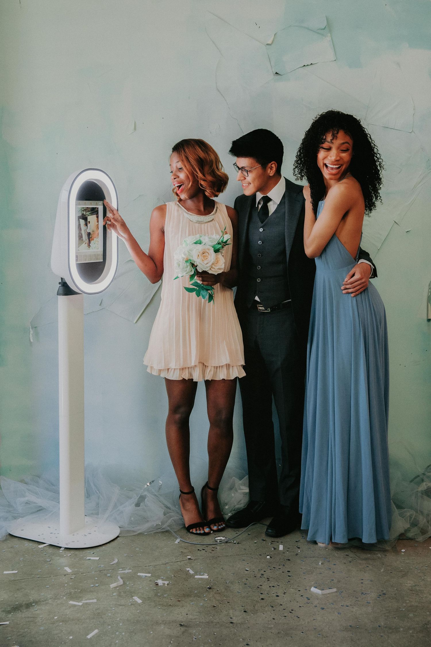 A group of wedding guests posing and laughing together next to a photo booth kiosk against a light blue wall.