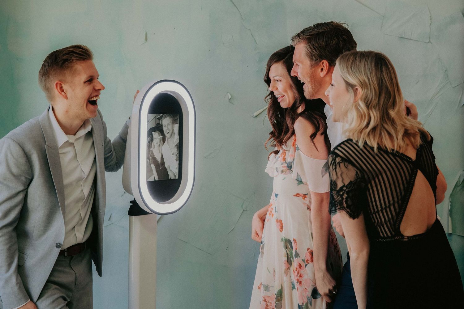 A group of people laugh and smile together while posing in front of a mint green wall for candid party photos.