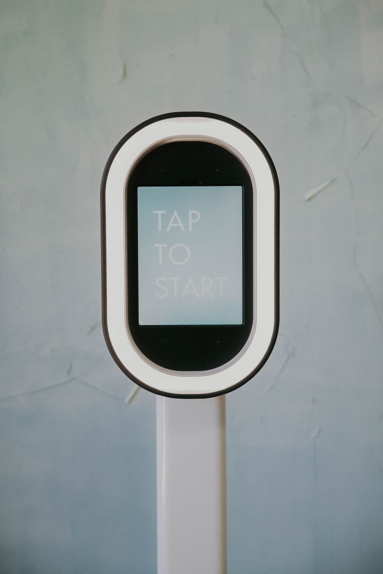 A modern white digital kiosk display showing "TAP TO START" text on a black screen against a gray background.