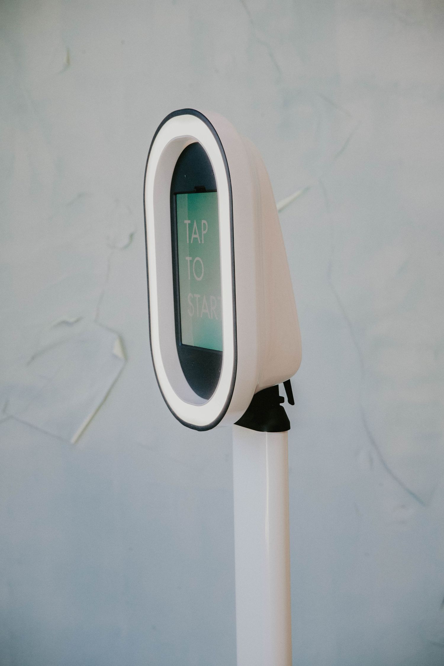 Modern white digital payment terminal with illuminated screen mounted on a pole against light grey wall.