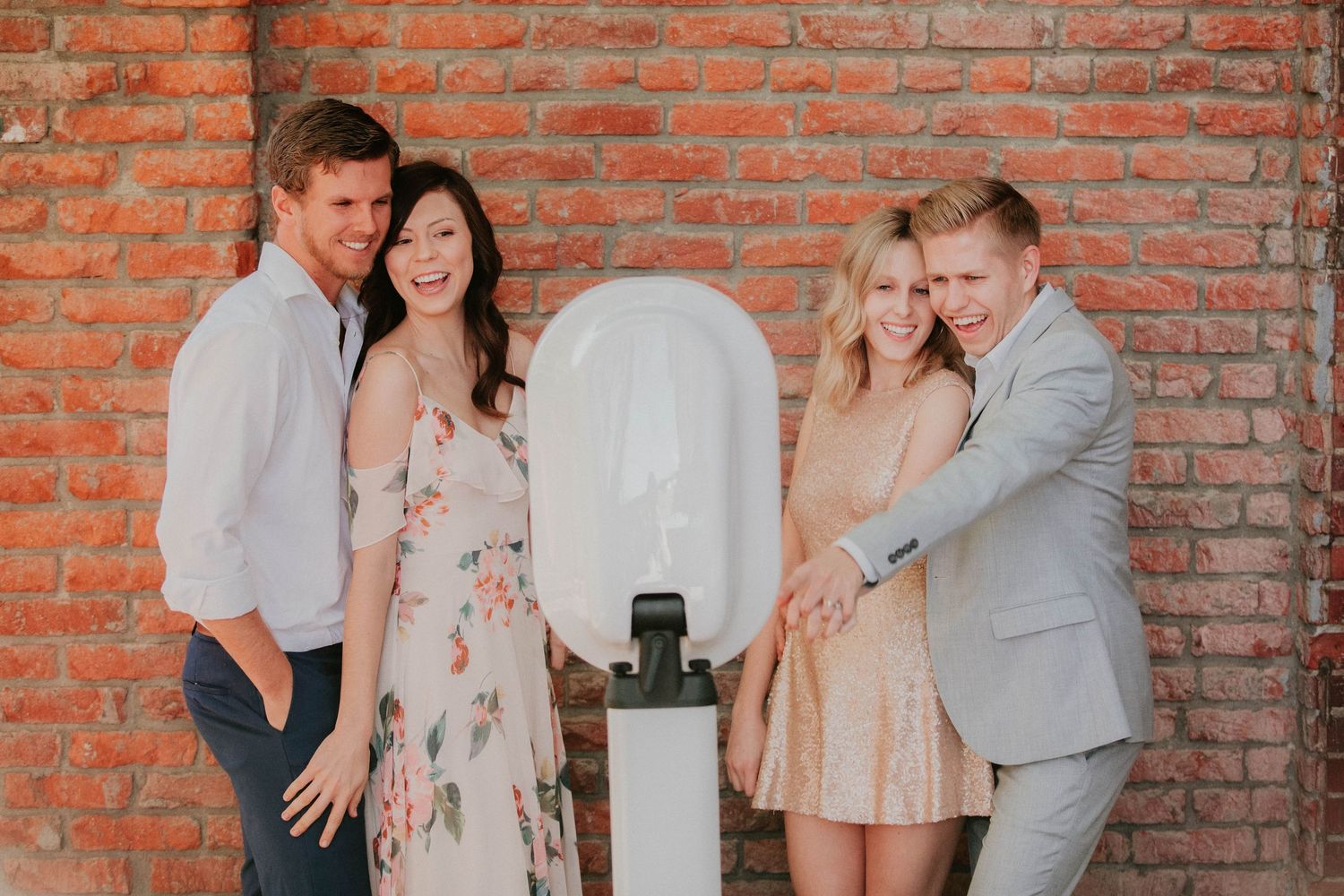 Two couples pose together against a rustic brick wall with a white architectural element between them.