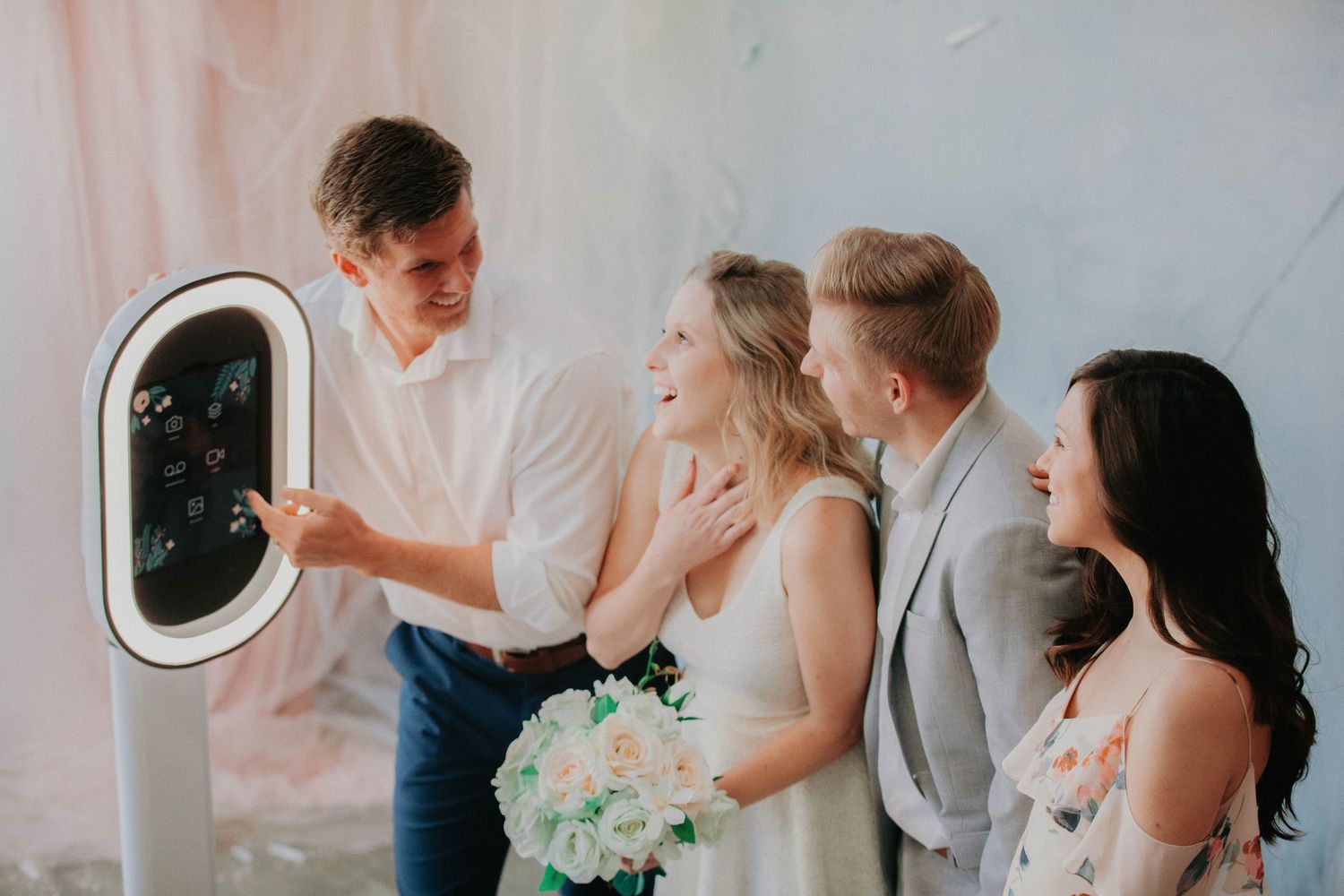 People gather around a photo booth machine at a wedding celebration with blue bouquet and light colored backdrop.