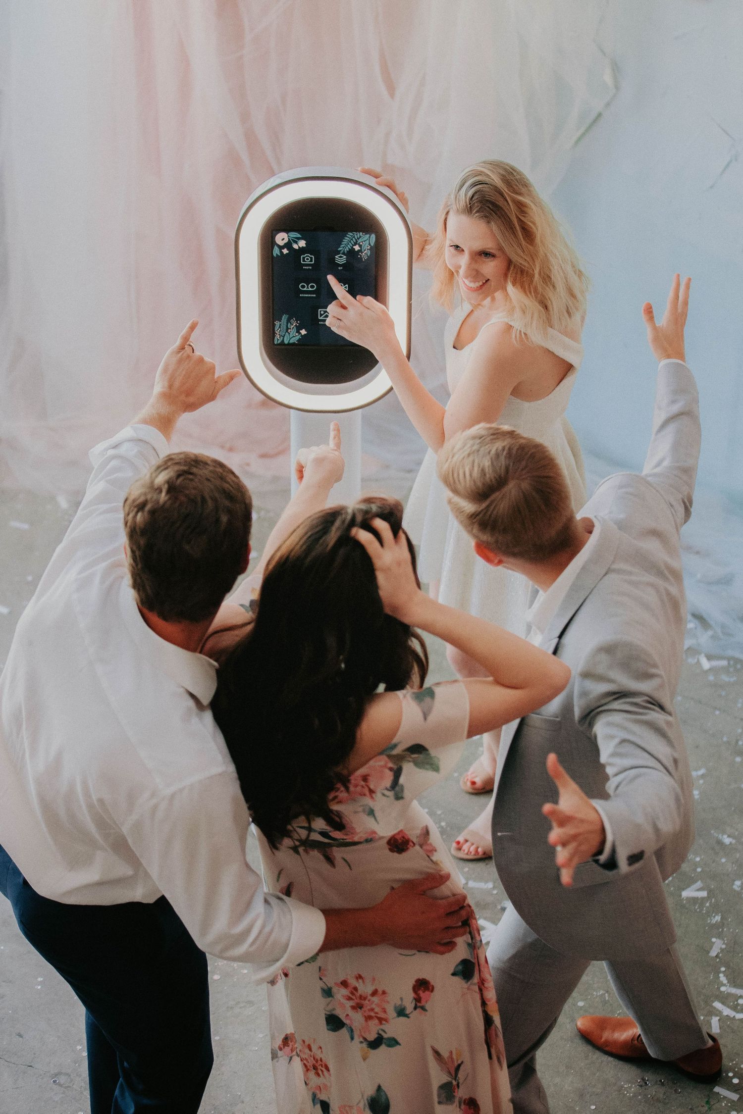 A group of friends excitedly gather around a photo booth touchscreen display at a social event.
