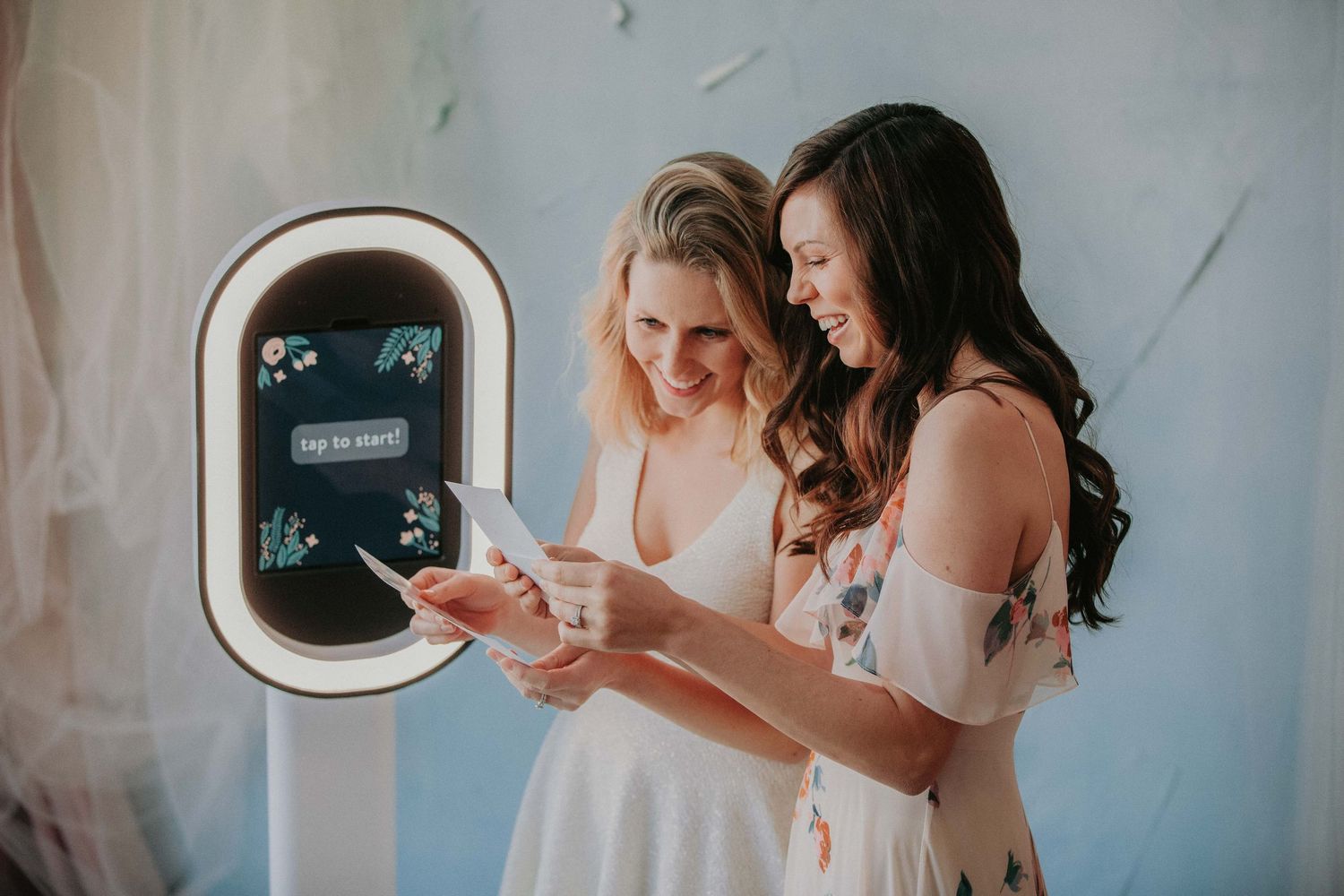 Two women in white dresses interact with a digital photo booth at a social event.