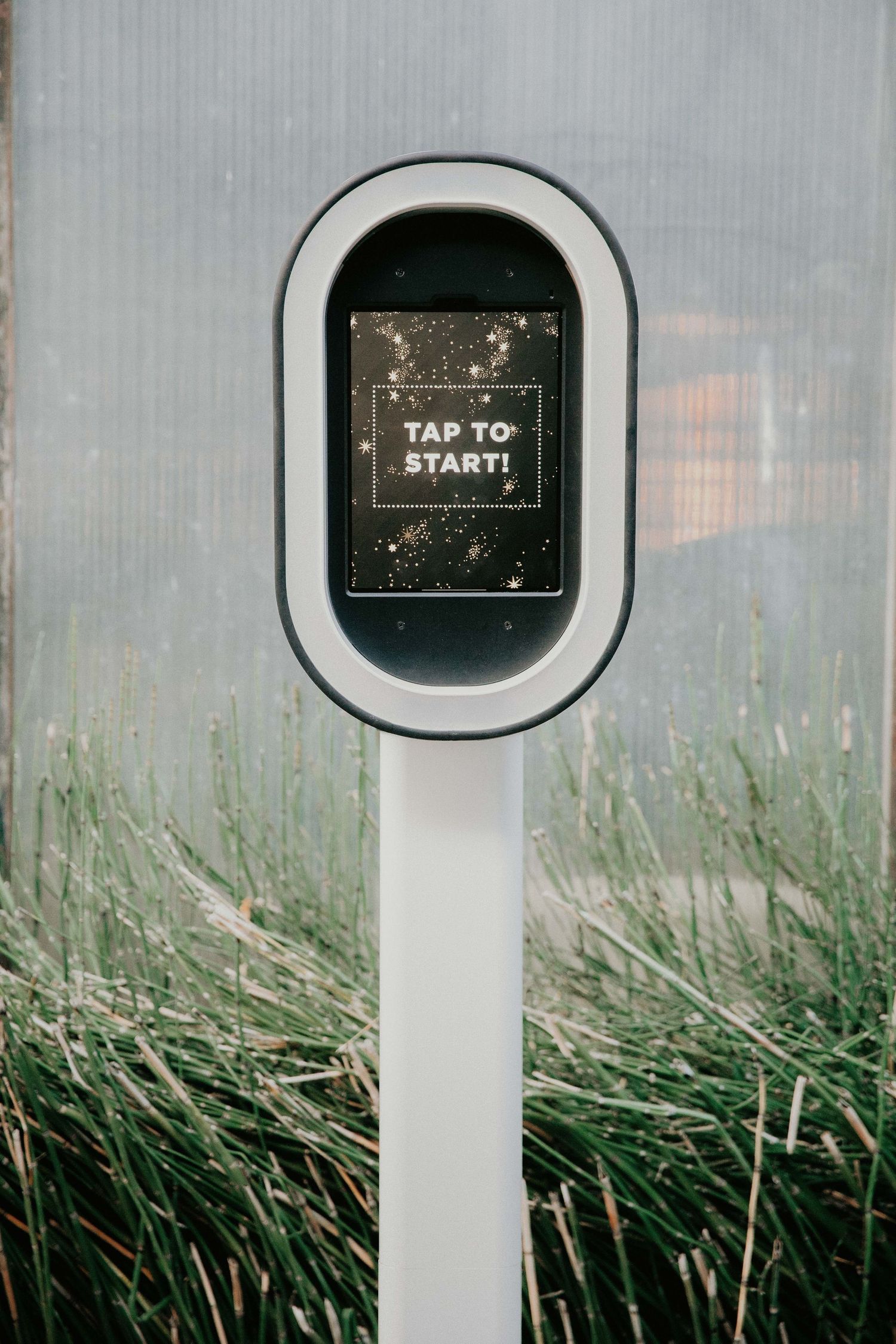 A white pedestal display screen showing 'TAP TO START' text against tall grass in the background.