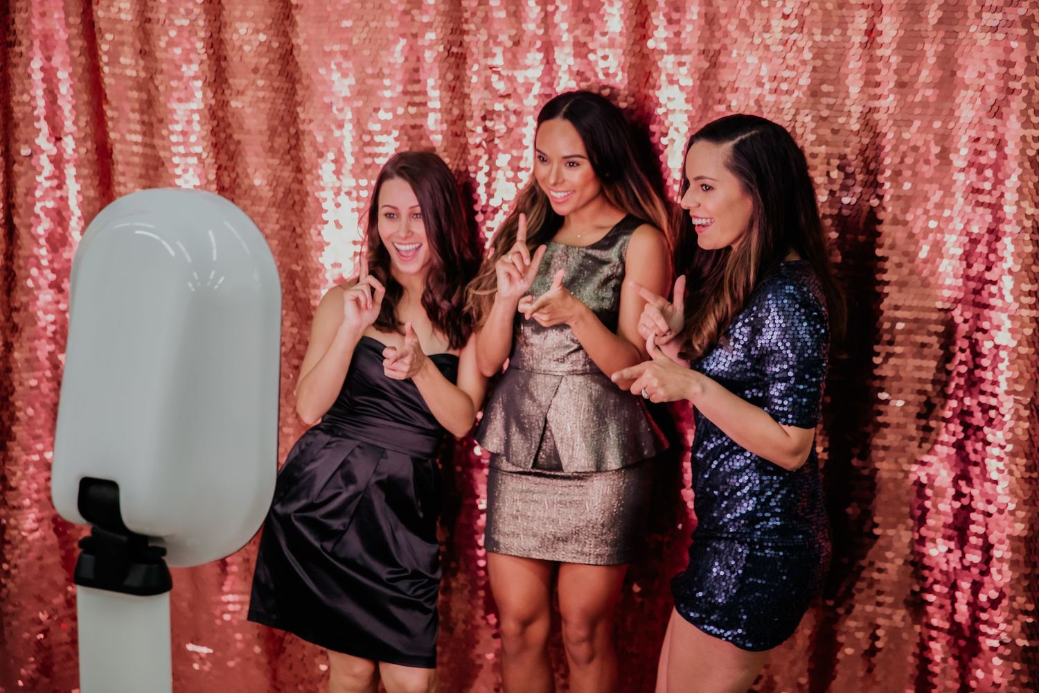 Three friends pose and laugh together in front of a pink sequin backdrop next to a photo booth setup.
