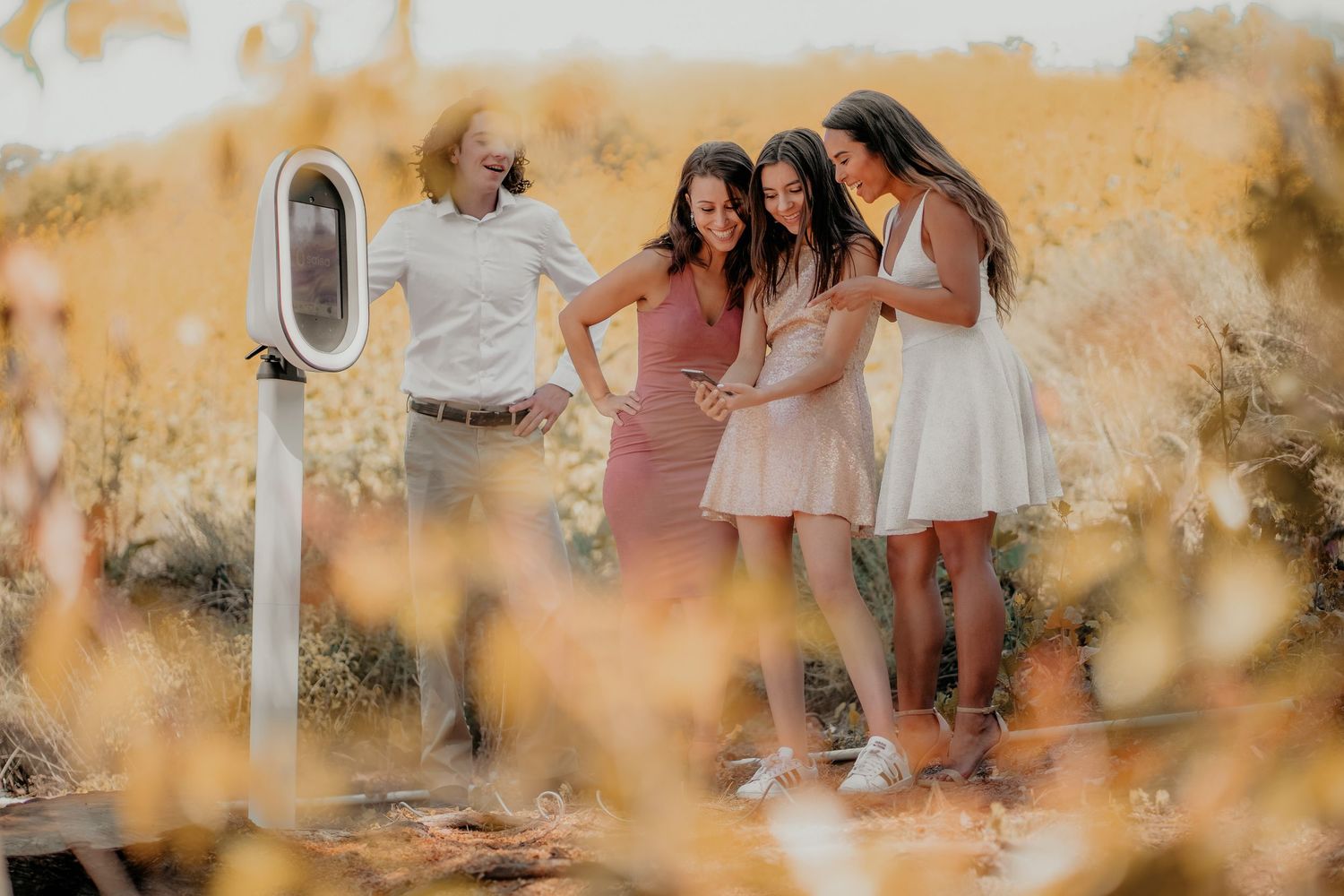 Friends in summer dresses gather around a machine in a golden field at sunset, sharing a joyful moment together.