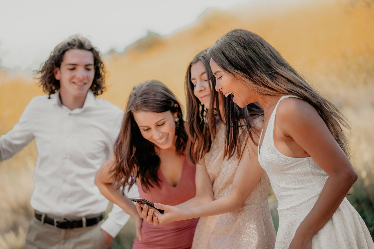 A group of friends stand close together looking down at something while laughing in a sun-lit outdoor setting.