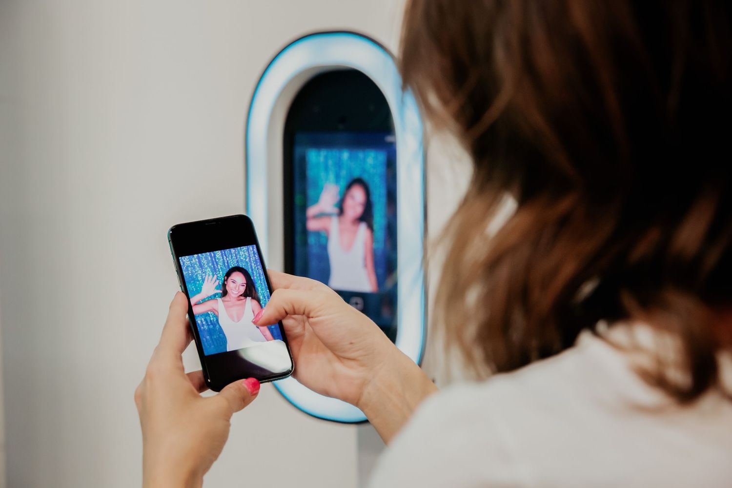 Someone holds a smartphone to take a photo of their reflection in an illuminated smart mirror display.
