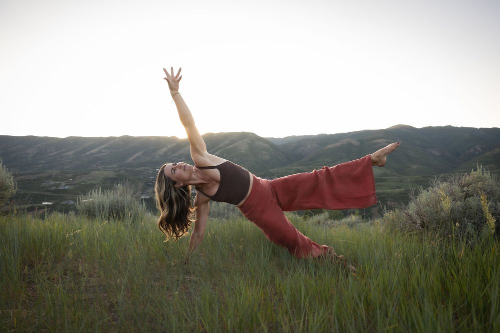 A portaits of a woman practicing yoga in a serene mountain landscape with tall grass and rolling hills in the background.