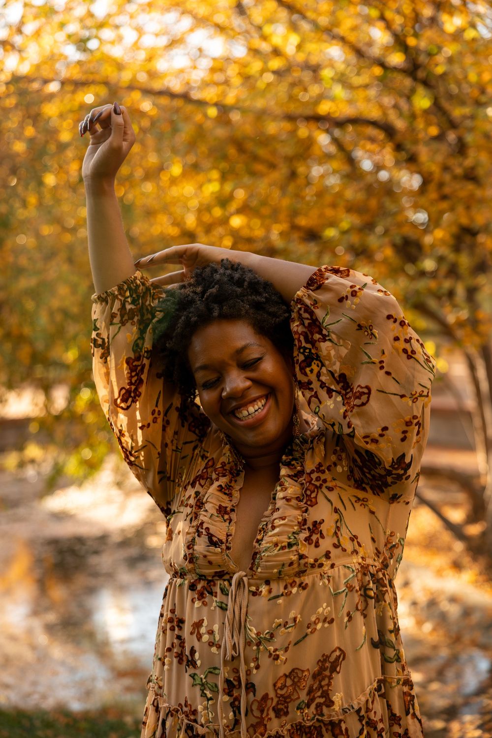 A portrait of a person dancing joyfully in a flowy floral dress against a backdrop of golden autumn trees during sunset.