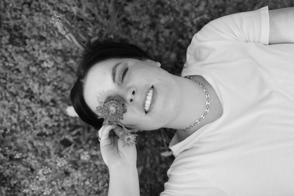 Black and white portrait of woman lying on grass with closed eyes and a peaceful smile wearing a white top, holding a flower over one eye.