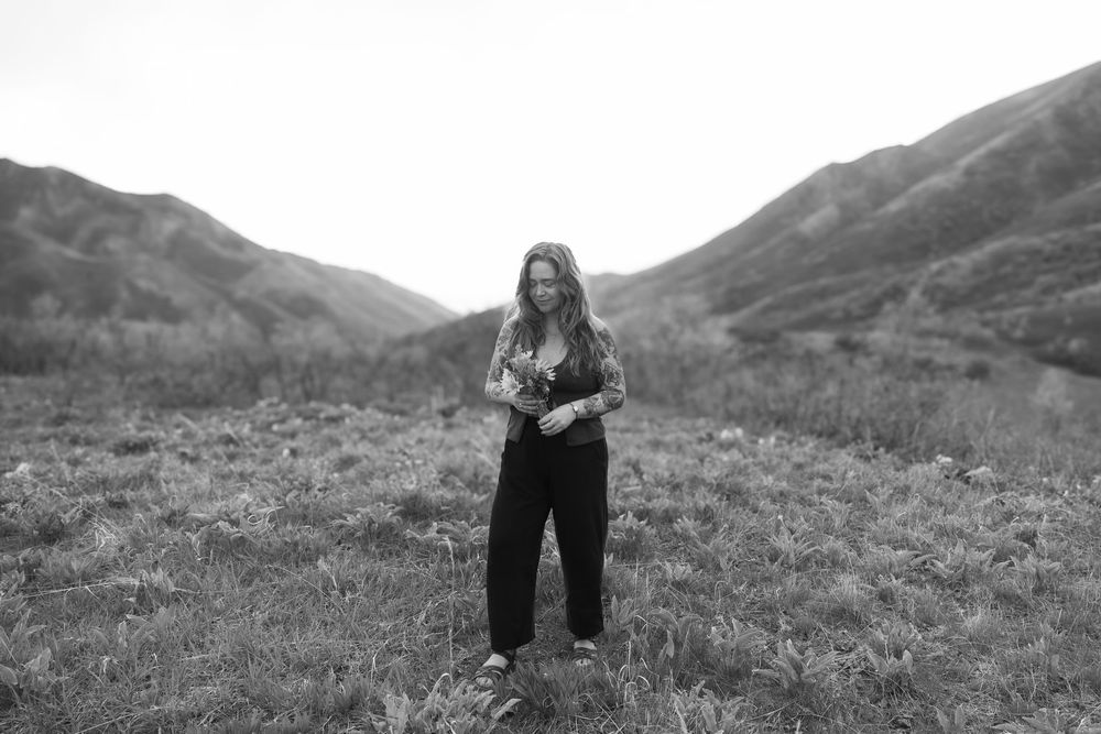 A woman holding a bouquet stands on a grassy hillside overlooking a mountainous landscape in black and white.