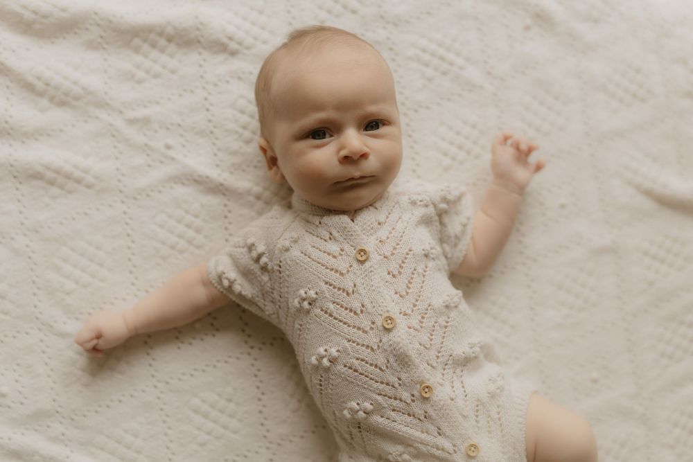 A baby wearing a white knit sweater lies on cream-colored bedding while stretching their arms outward.