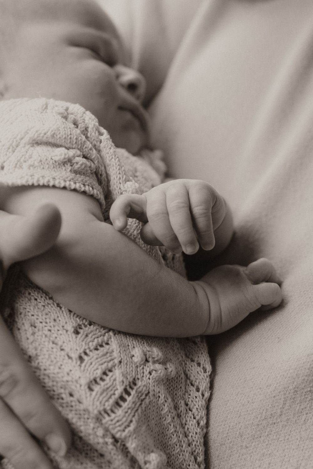 Close-up black and white photo of a sleeping baby in a knit sweater showing tiny curled fingers and peaceful expression.