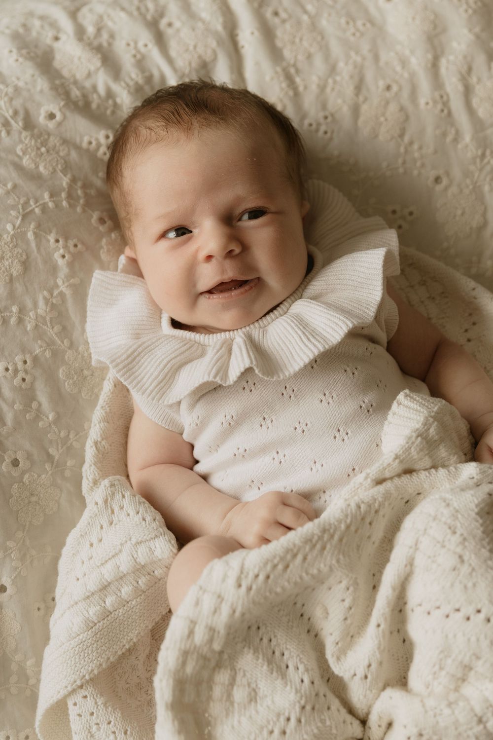 A baby wearing a white ruffled collar dress lies on a textured cream-colored blanket with a gentle smile.