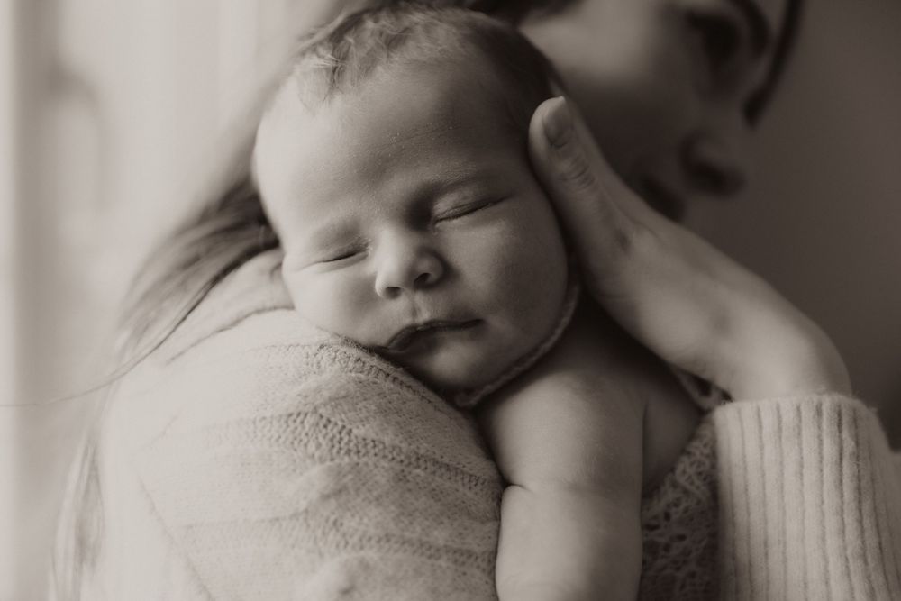 A sleeping newborn baby rests peacefully on a caregiver's shoulder in a soft black and white portrait.